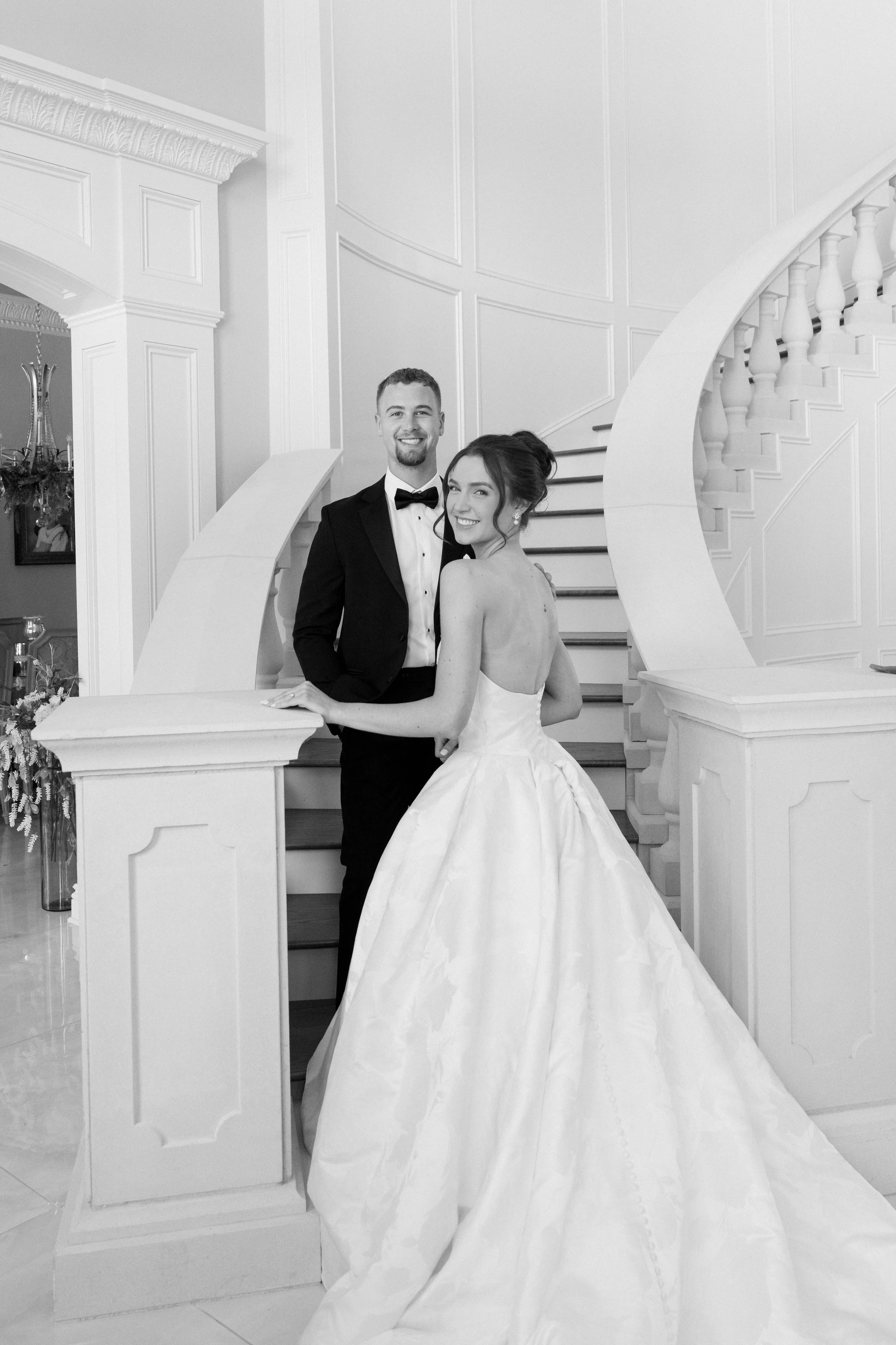 Luxury Wedding - Black and white photo of a bride and groom standing together inside a grand staircase with elegant white woodwork. The groom is wearing a tuxedo with a bowtie, and the bride is in a strapless wedding gown with her hair styled up.