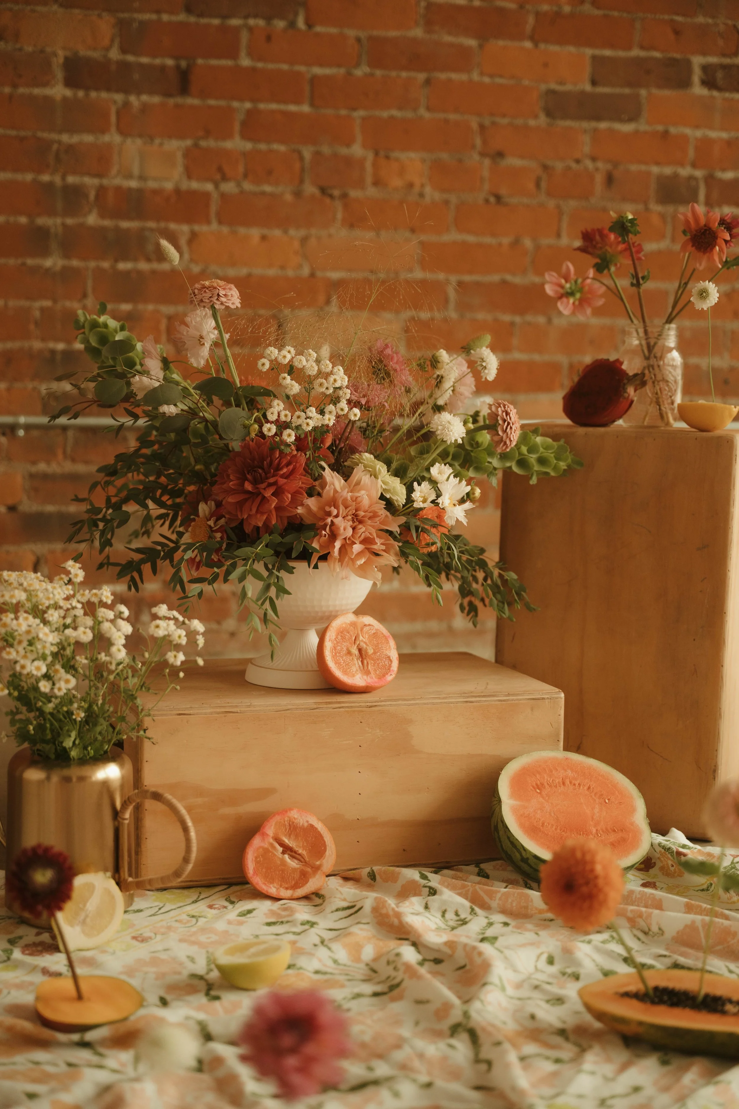 A floral arrangement with pink, white, and red flowers in a white vase, topped with a spider web, set on a wooden table against a brick wall background. There are sliced grapefruits and lemons, and other decorative items on the table.