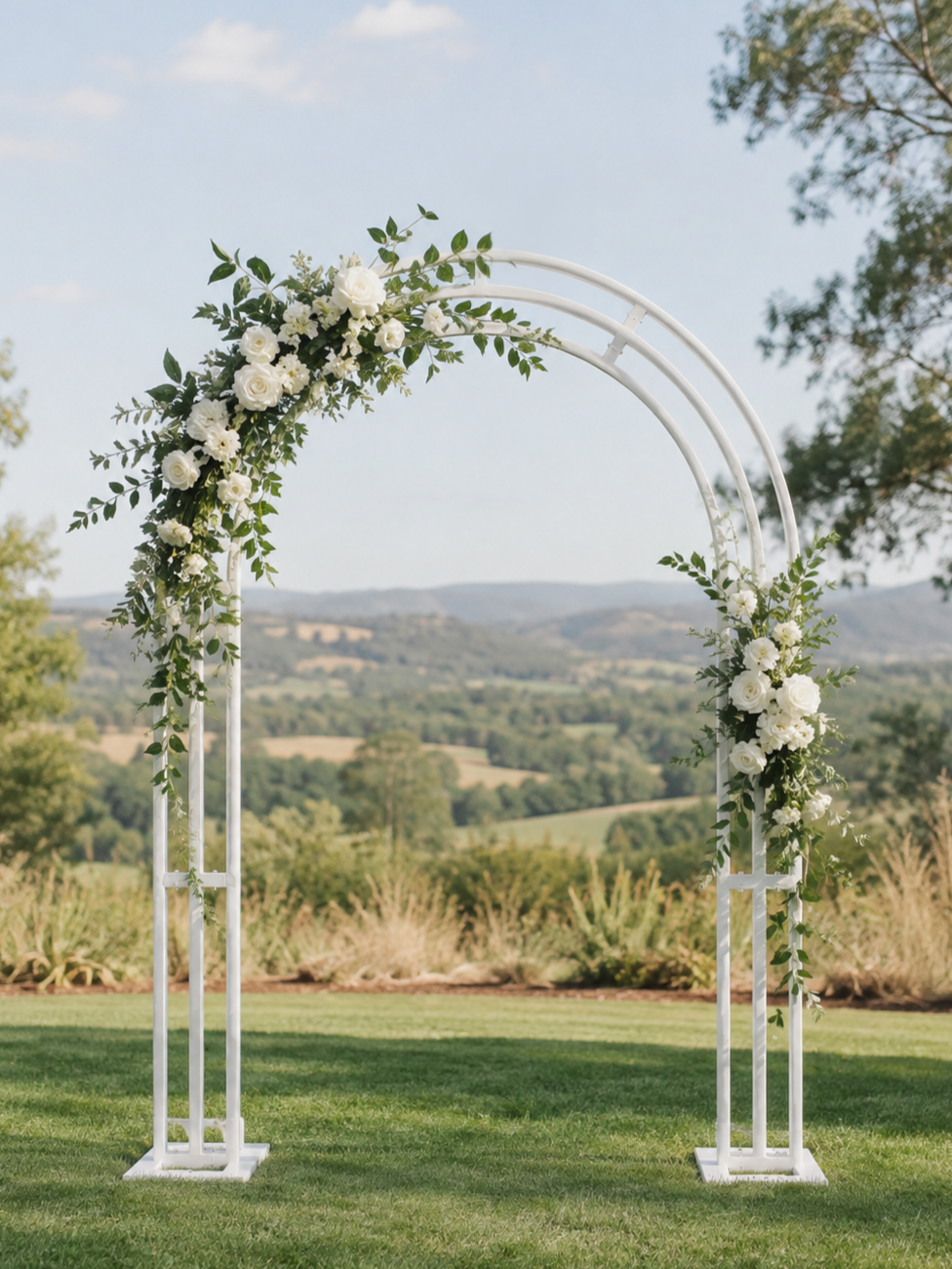 White wedding arch decorated with white roses and greenery on a grassy field with a scenic rural landscape in the background.
