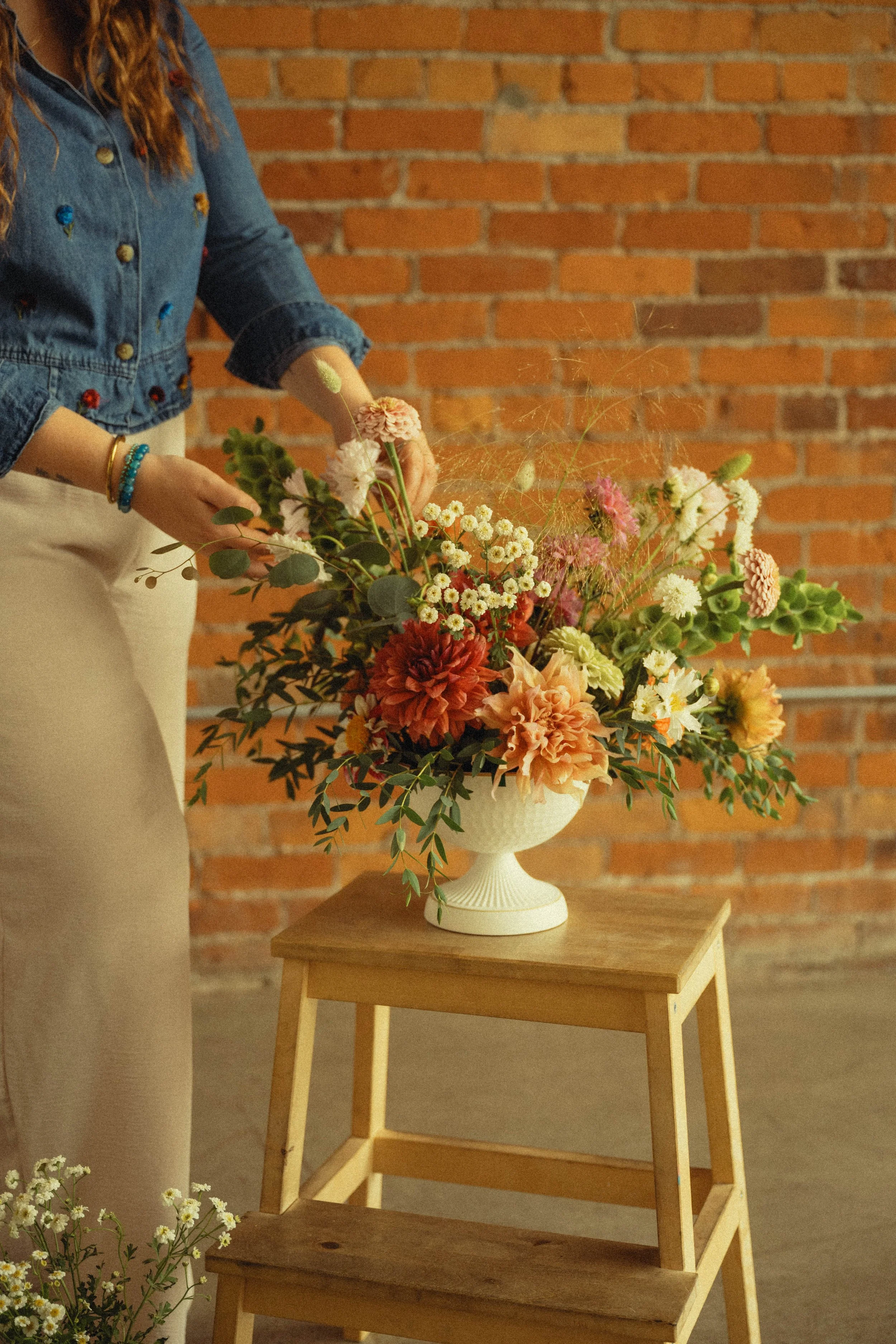Woman arranging a bouquet of mixed flowers on a wooden stool against a brick wall.