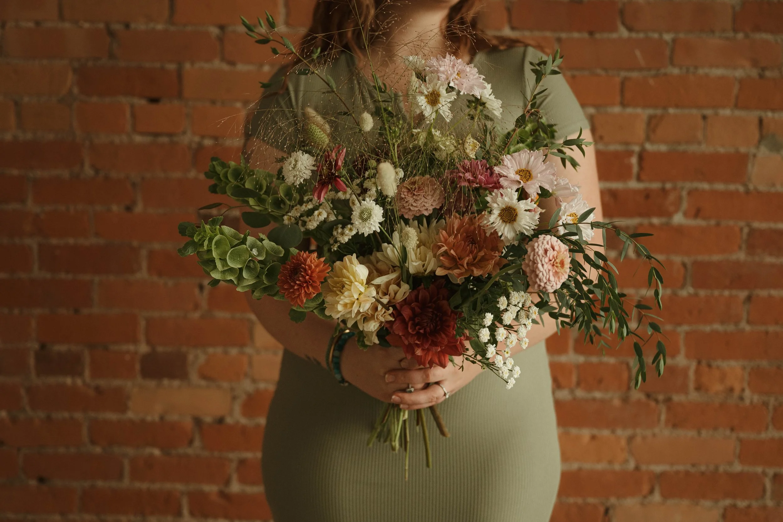 Person standing against a brick wall, holding a large bouquet of mixed flowers.