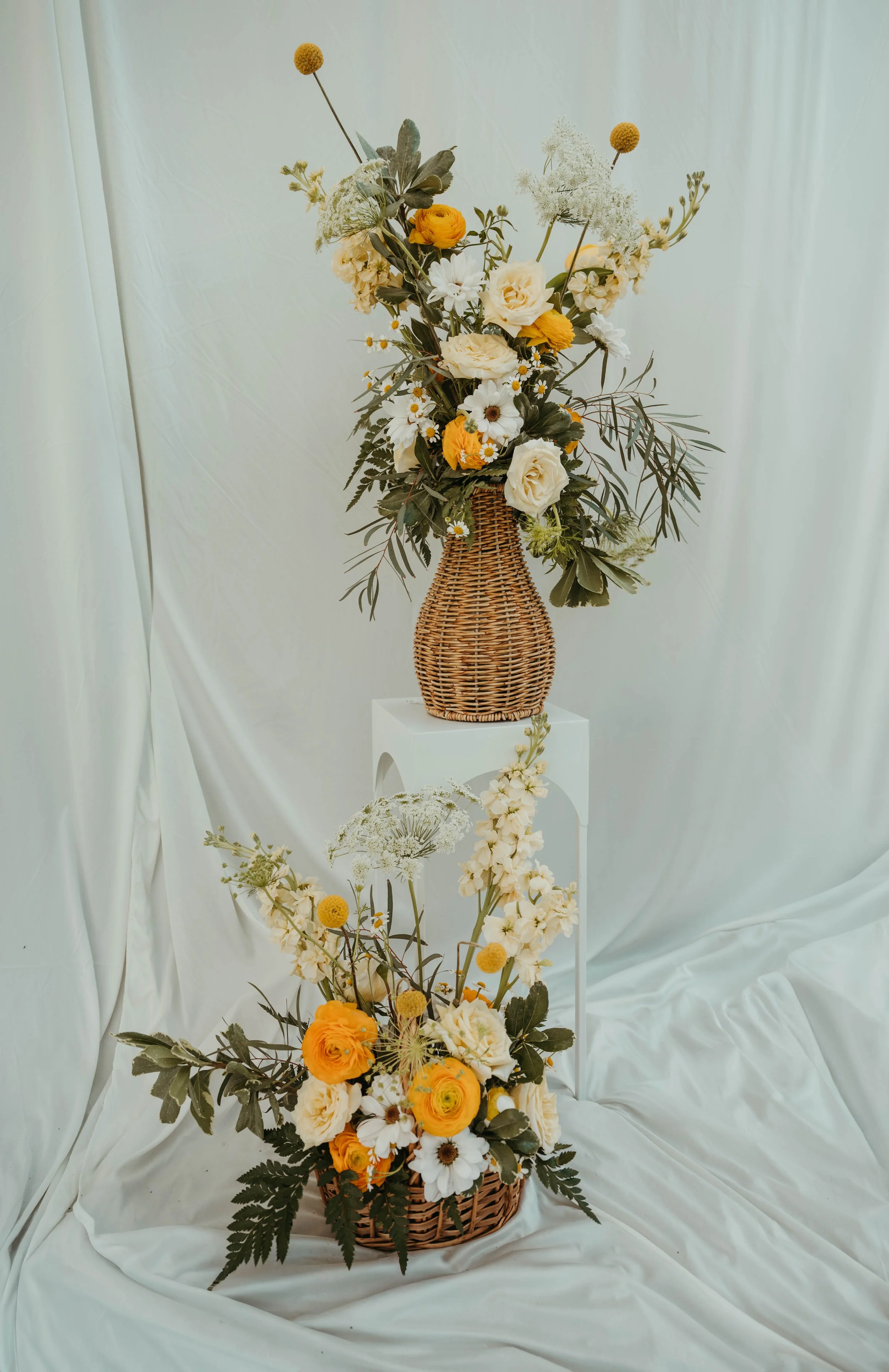 A tall wicker vase with a bouquet of flowers, including white, yellow, and orange blooms, arranged against a pale fabric backdrop. The arrangement is displayed on a white pedestal, with additional flowers and greenery at the base.