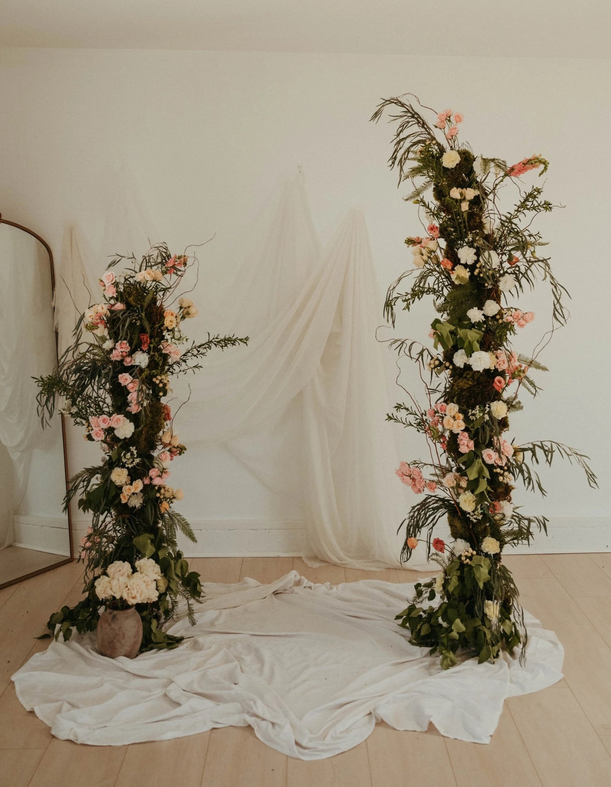 Two large floral arrangements with pink and white flowers and greenery on dark stands, positioned in front of a white fabric backdrop, with a mirror on the left and white fabric draped on the floor.