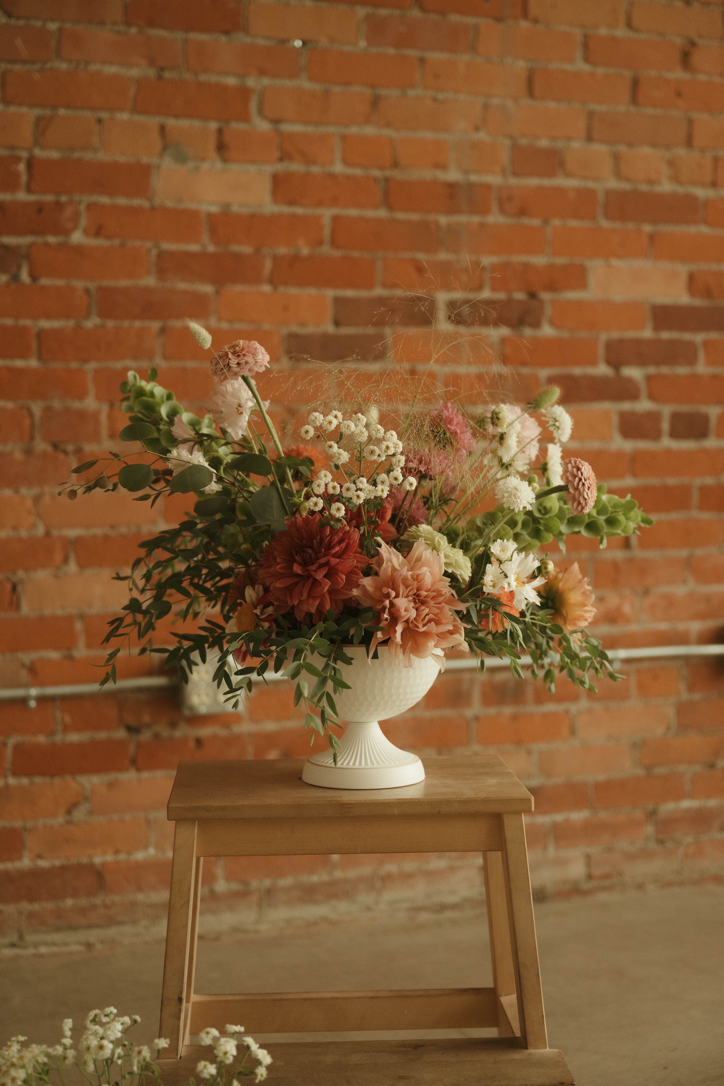 A floral arrangement in a white pedestal vase on a wooden table against a brick wall.