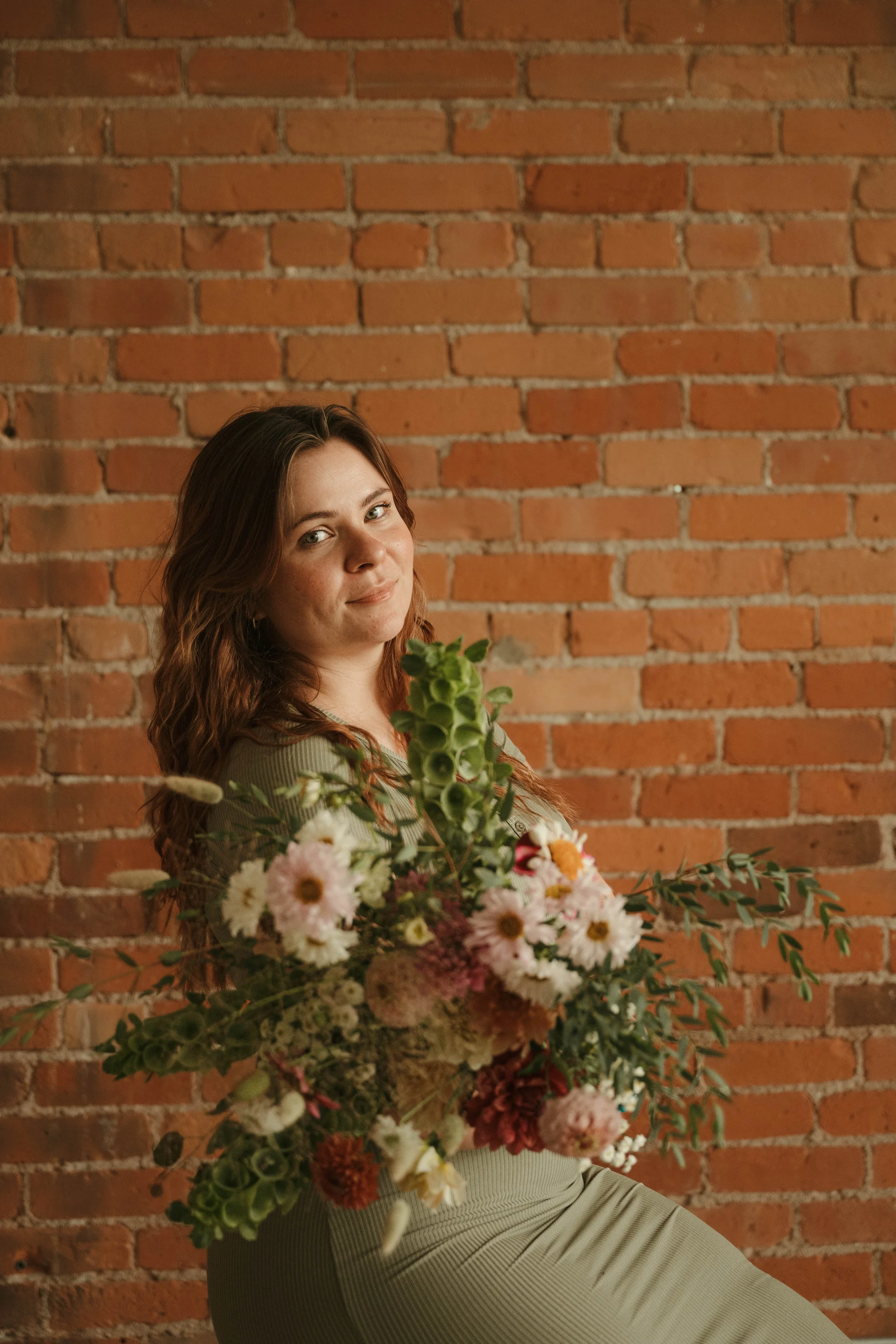 A woman with long brown hair holding a large bouquet of pink, white, and orange flowers, standing in front of a red brick wall.