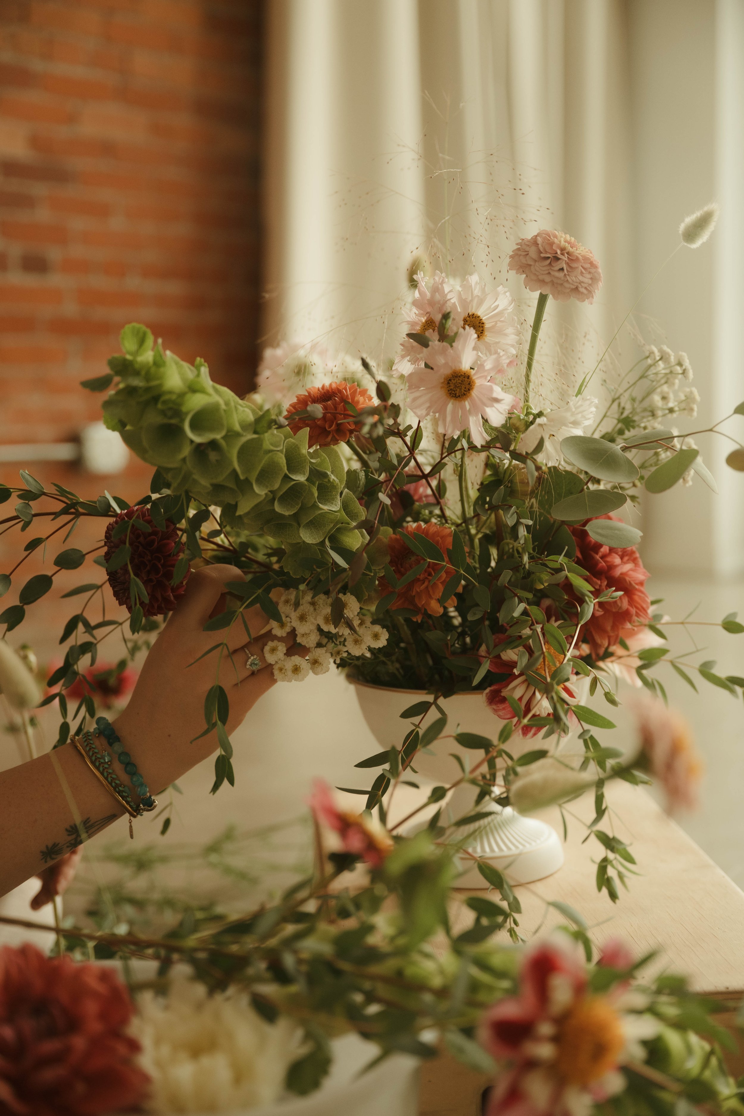 A hand holding a bouquet of mixed flowers, including pink, white, and orange blossoms, with greenery and delicate spider webs, set in a room with a brick wall and curtains.