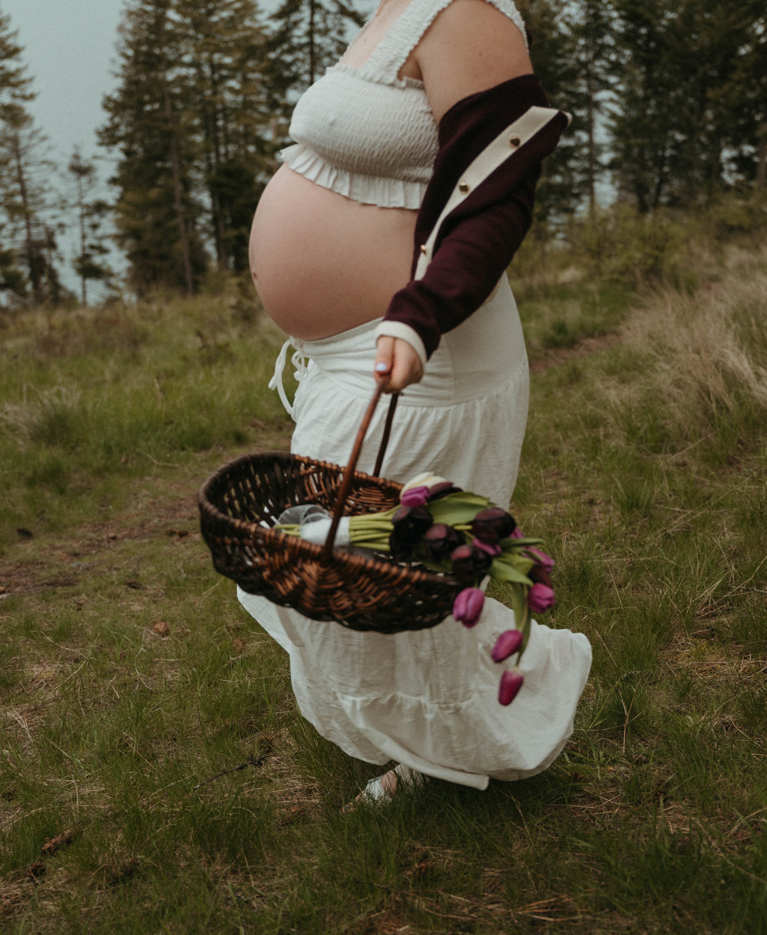 A pregnant woman wearing a white dress and a sleeveless top holds a basket of pink tulips in an outdoor grassy area with trees in the background.