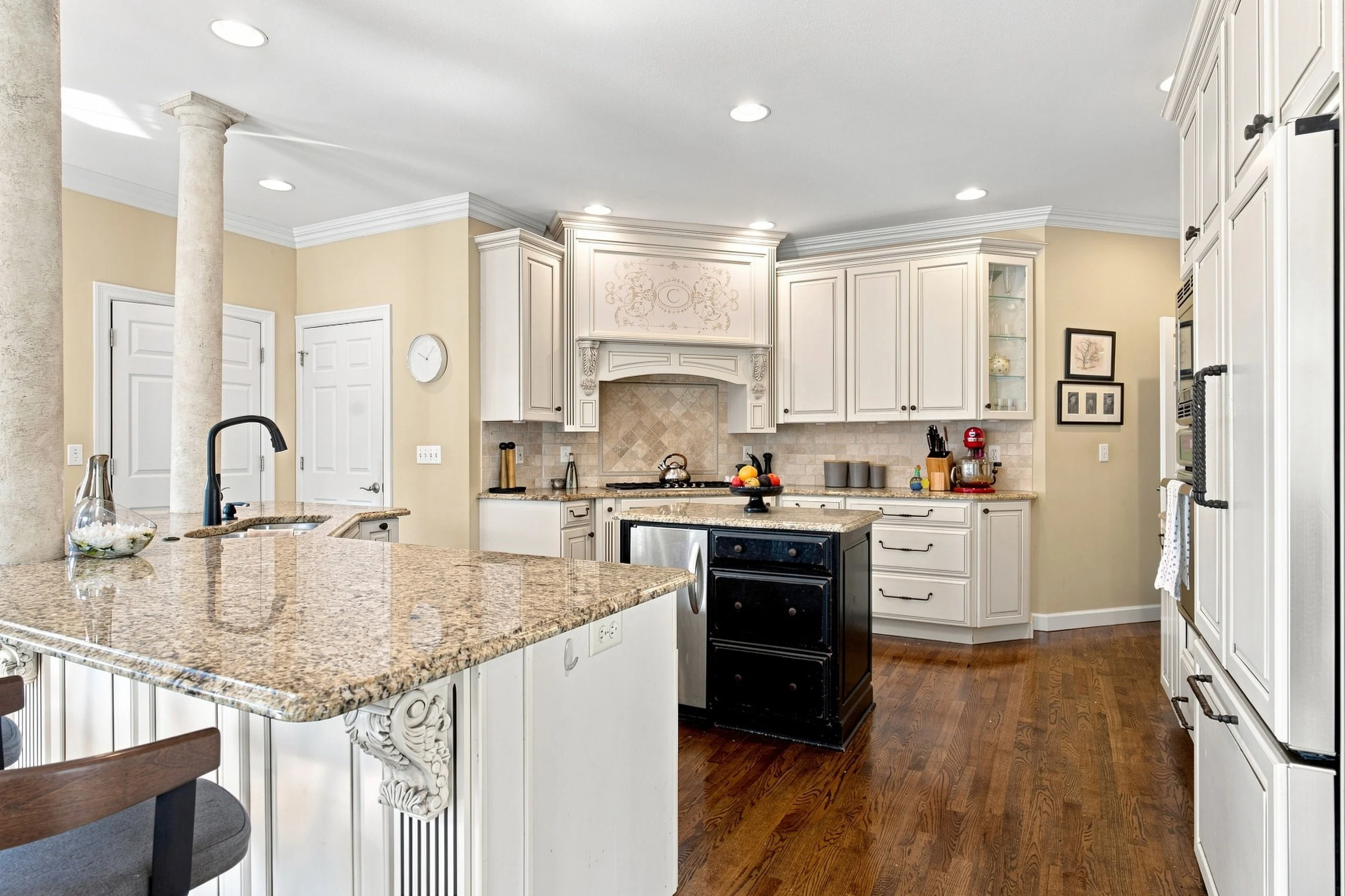 A spacious kitchen with white cabinets, granite countertops, and wooden flooring. Features a black oven, a kitchen island with a sink, and various small appliances and decorative items on the counters.
