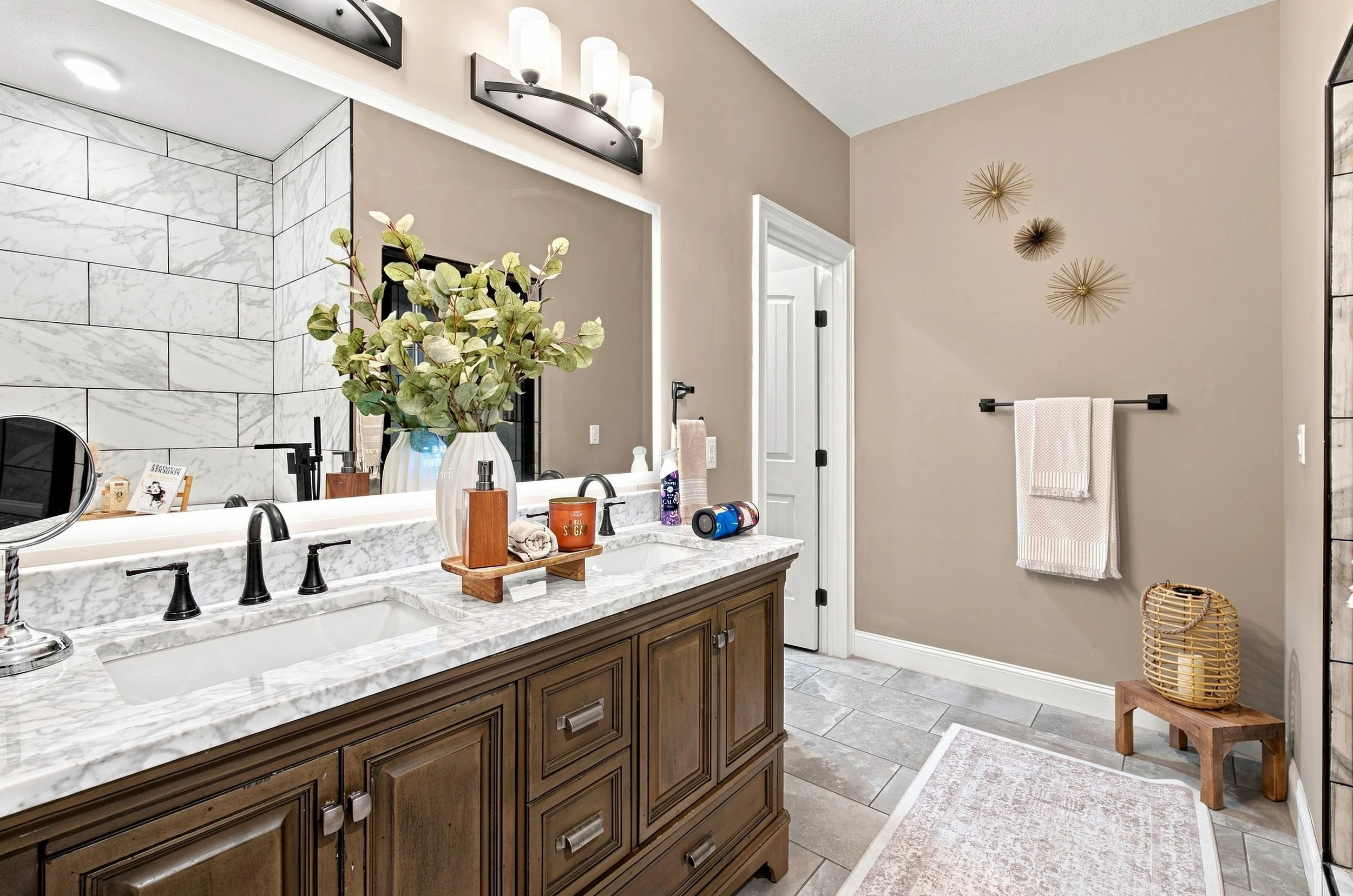 Bathroom with beige walls, a large mirror above a marble countertop with two sinks, and a wooden vanity cabinet. Decor includes a green plant in a white vase, hand soap, toothbrush holder, and personal items. There is a towel hanging on a black towel