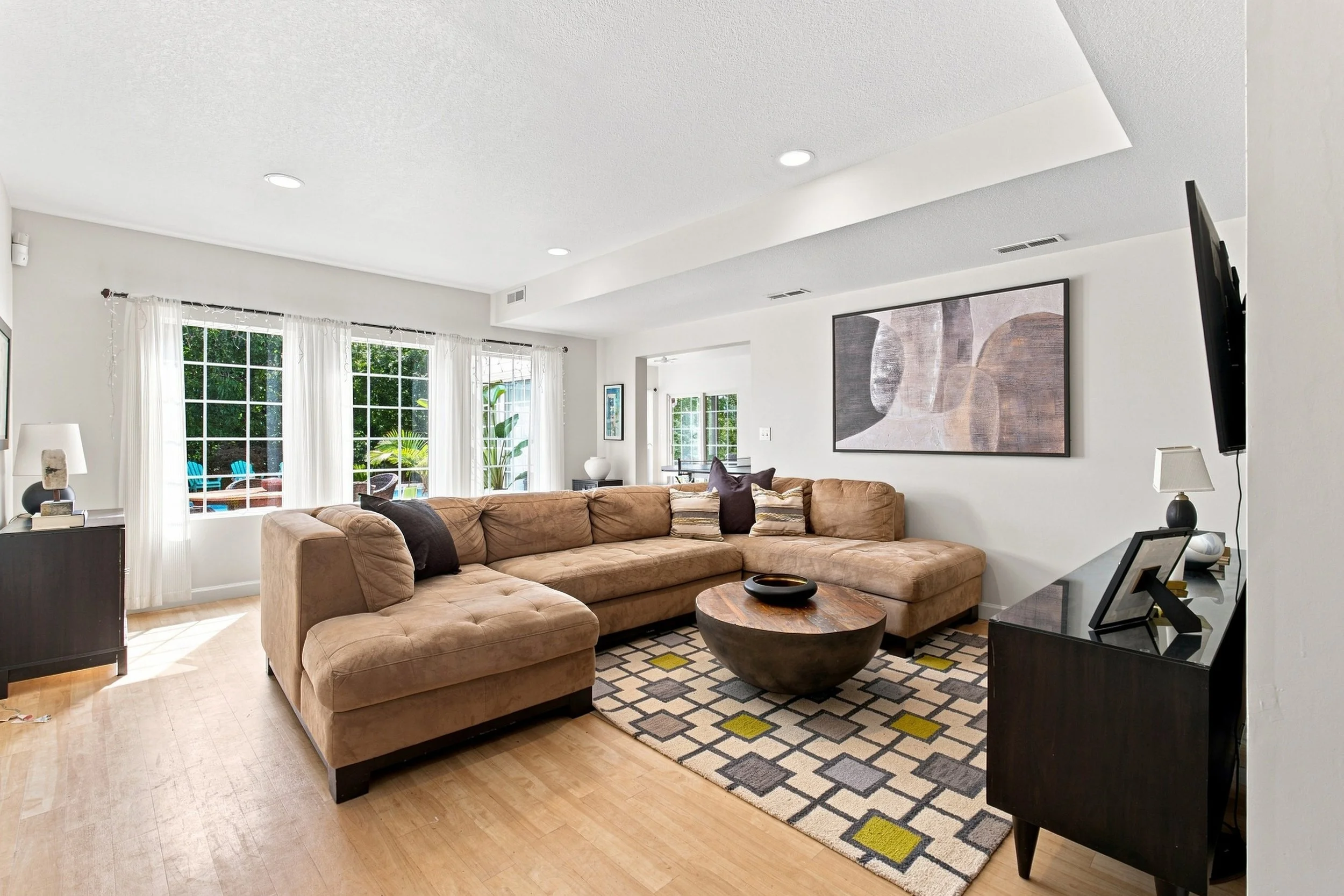 A living room with a beige sectional sofa, a patterned area rug, a black side table with picture frames, and large windows with white curtains letting in natural light.