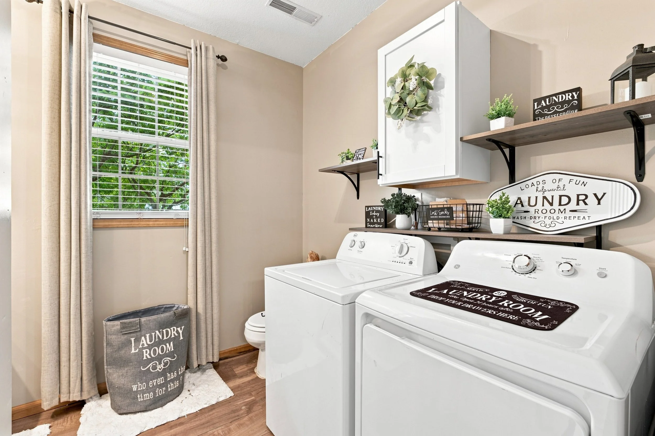 A laundry room with beige walls, a window with blinds, two white top-loading washing machines, and shelves with decor items and signs.