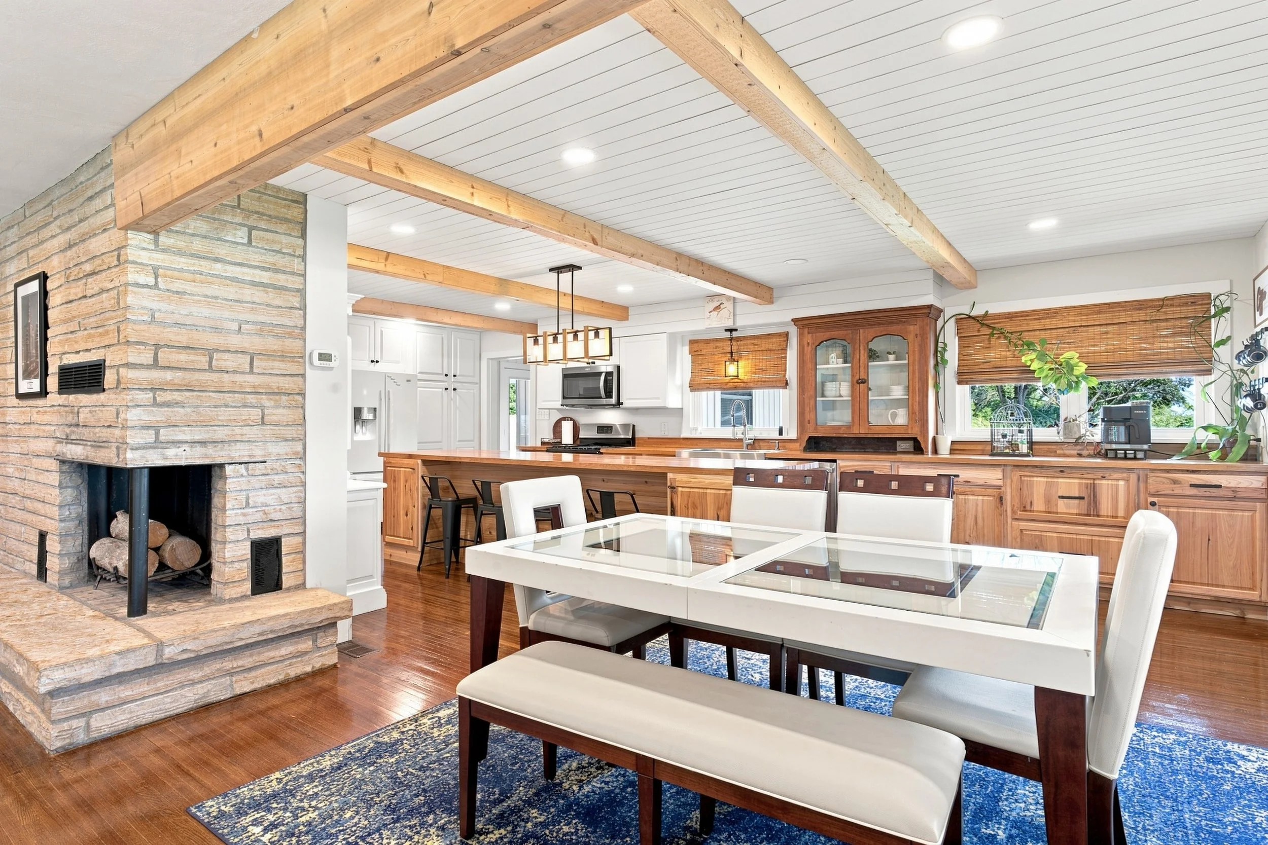 Open concept kitchen and dining room with exposed wood beams, white paneled ceiling, brick fireplace, hardwood floors, and large windows with blinds.