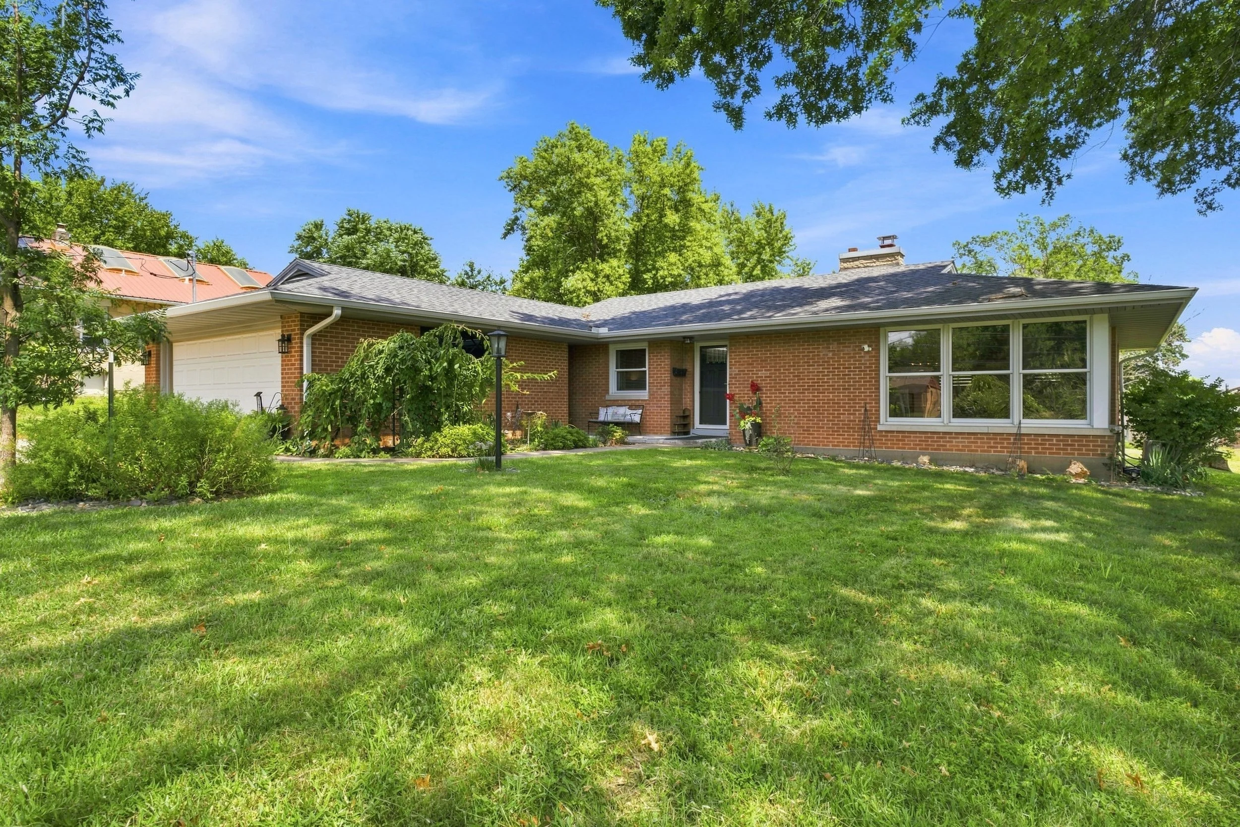 A brick house with large windows and a gray shingled roof, surrounded by green trees and a lawn under a partly cloudy blue sky.