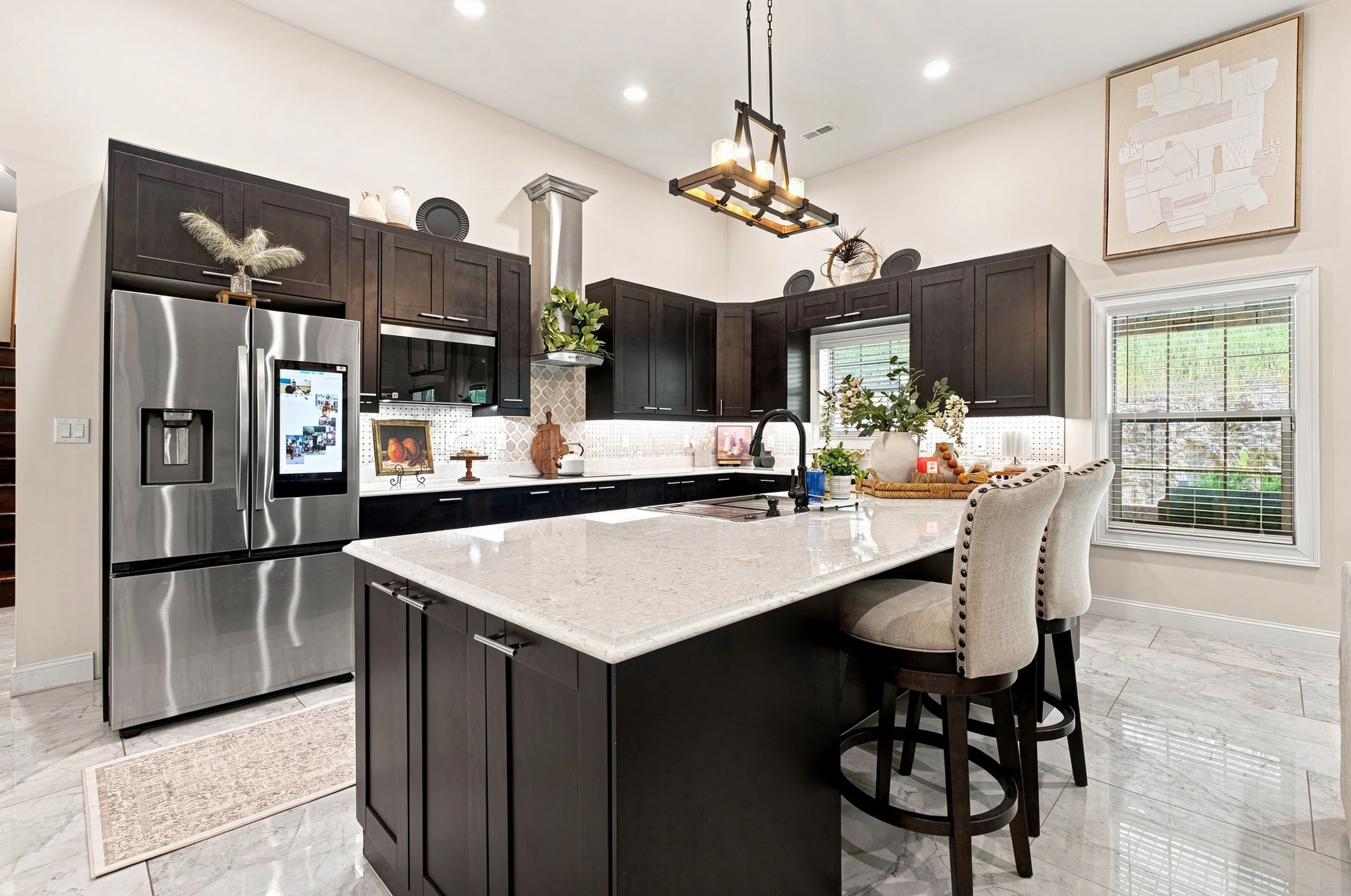Modern kitchen with dark wood cabinets, white marble island, stainless steel refrigerator, beige walls, and large window with blinds. Decor includes plants, vases, framed art, and a hanging light fixture.