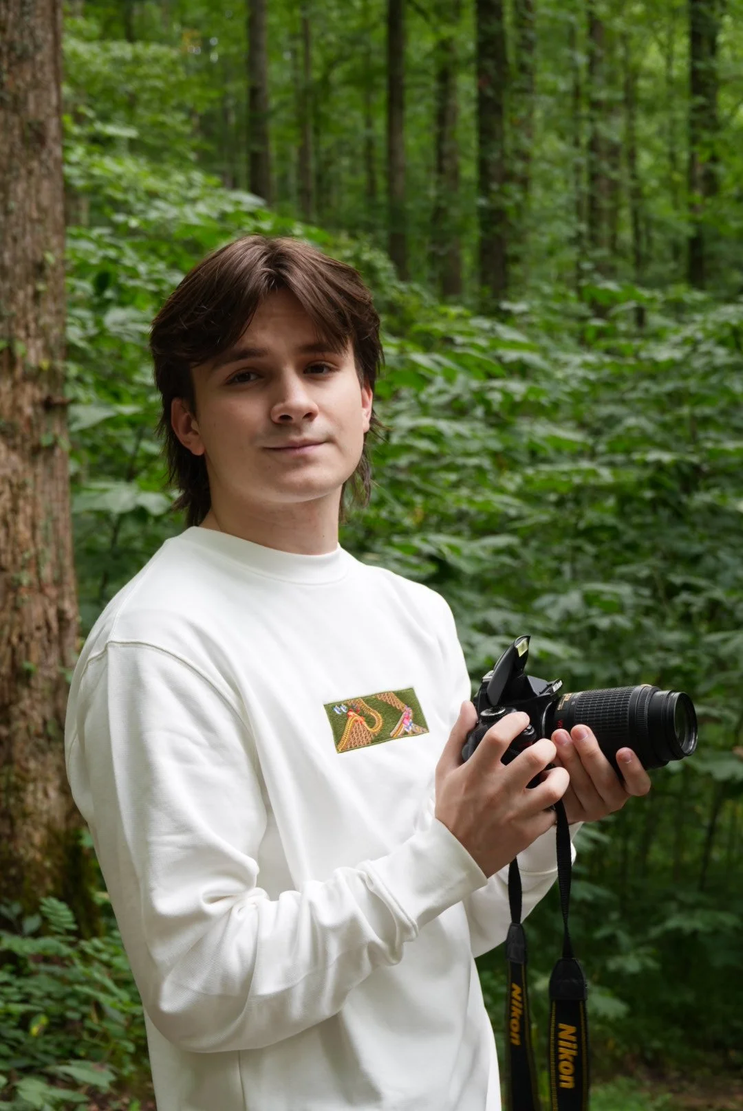 A young man stands in a wooded forest, holding a Nikon camera with a zoom lens, wearing a white sweatshirt with a colorful patch on the chest.