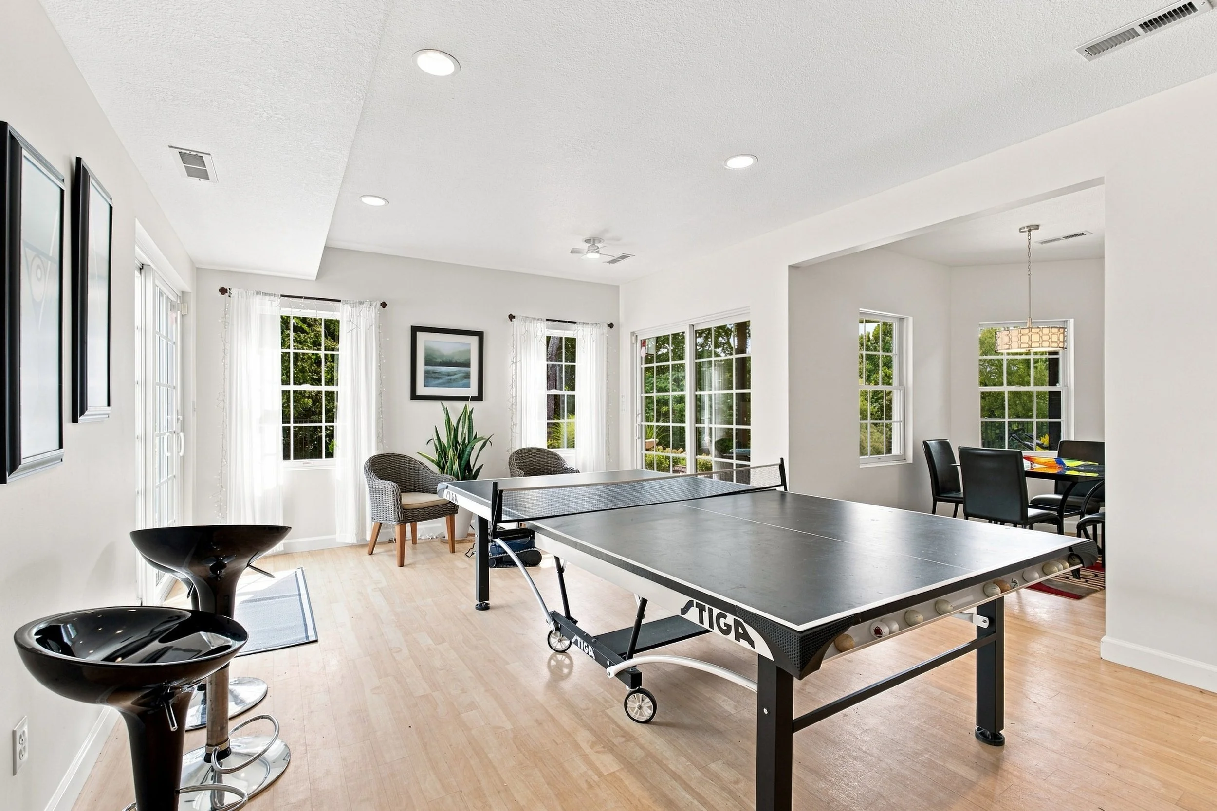 Bright living room with large windows, white walls, picture frames, a plant, and a ping pong table.