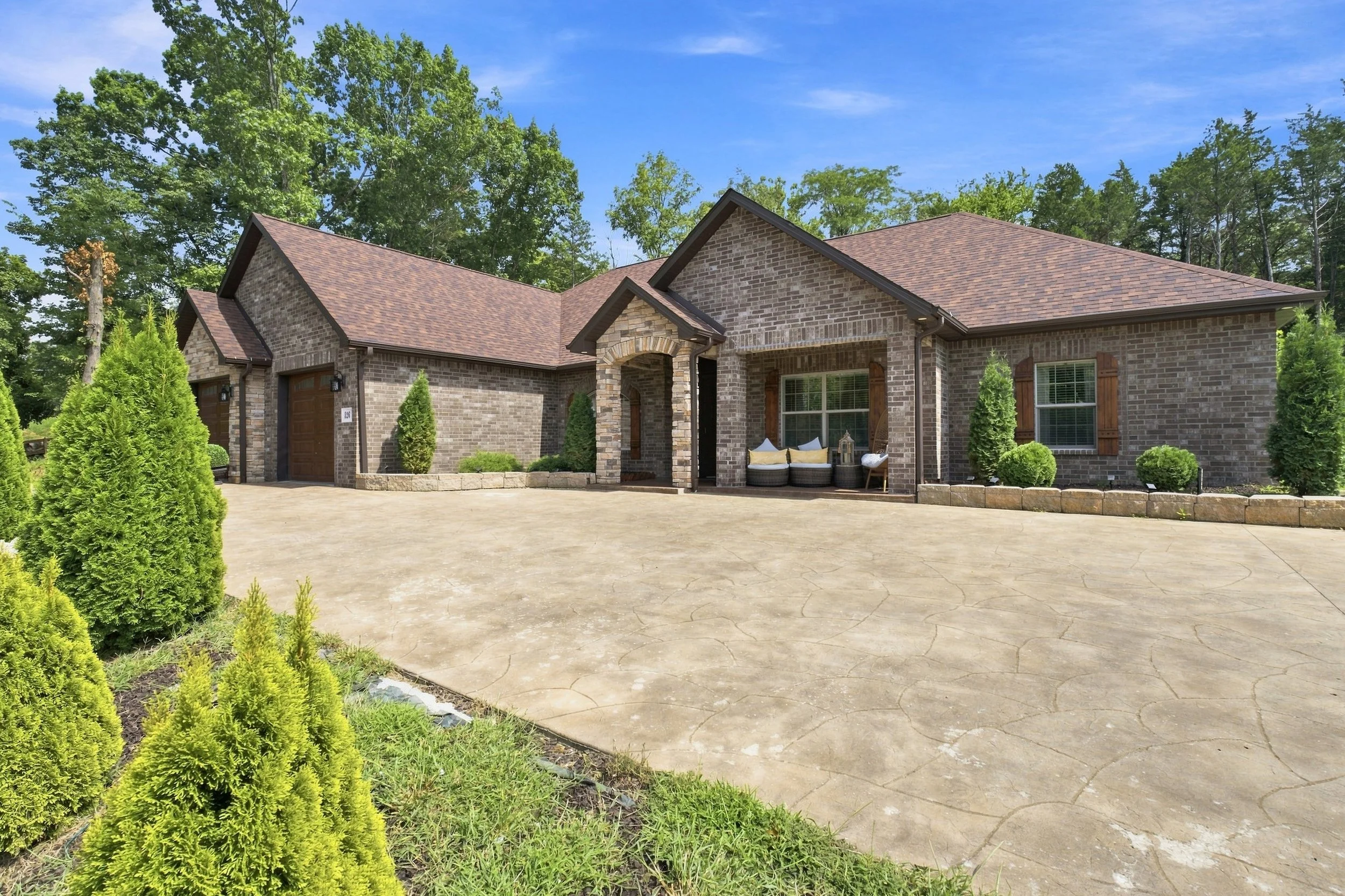 A brick house with a brown shingle roof, three garage doors, a covered porch with outdoor seating, and surrounding greenery, under a bright blue sky.