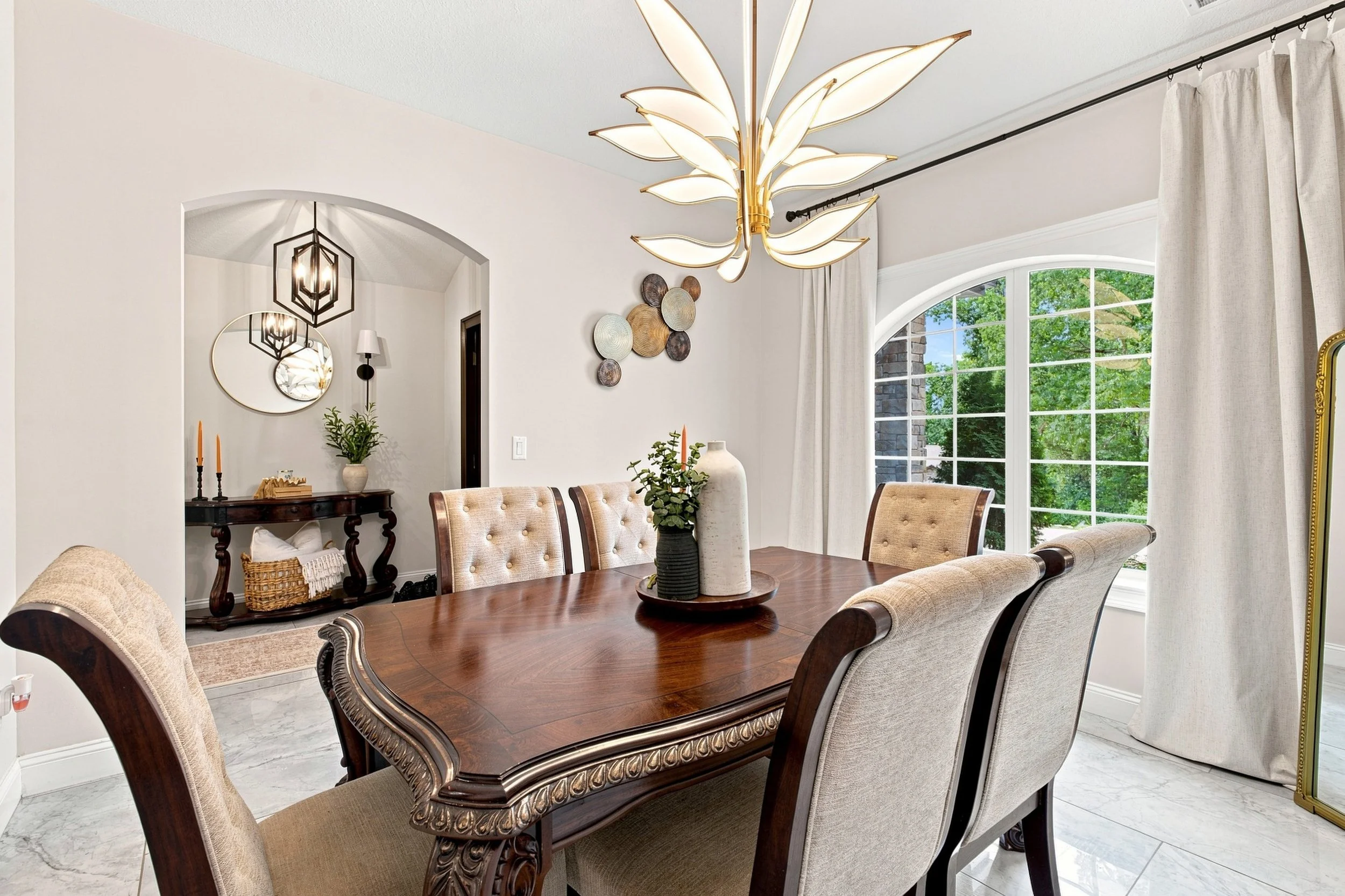 A dining room with a wooden table surrounded by beige upholstered chairs and decorated with potted plants and vases. Large windows with white curtains allow natural light, and a modern chandelier with leaf-shaped lights hangs above the table.
