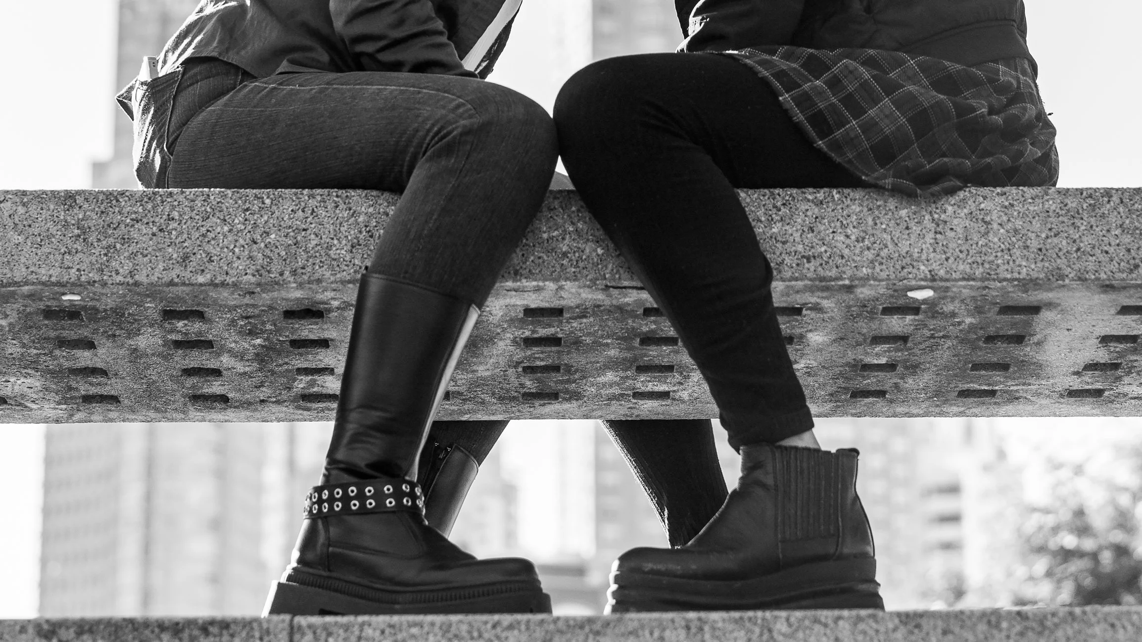 Two people sitting on a concrete ledge, one wearing rain boots and the other wearing shorter boots, with city buildings in the background.