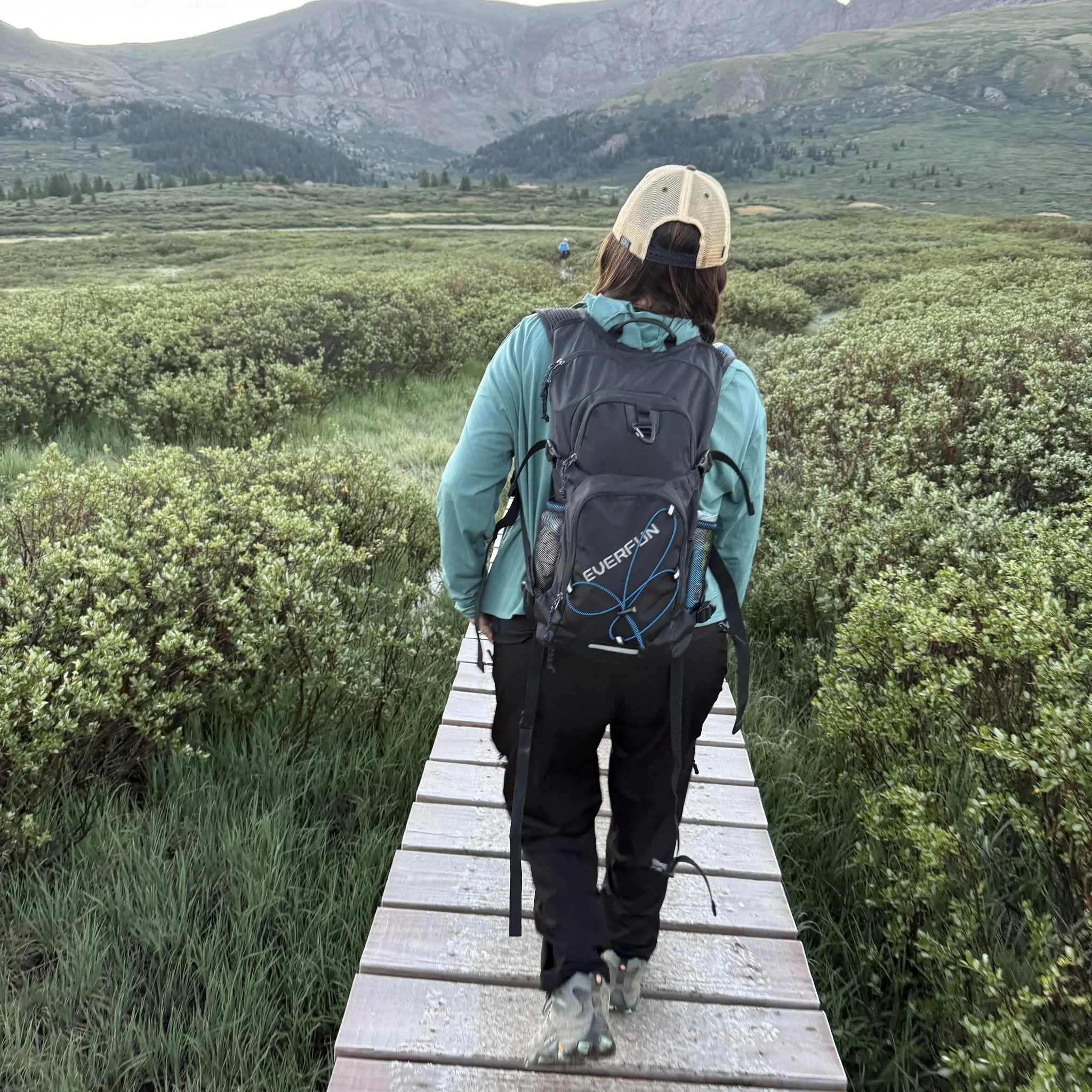 A person hiking in a mountainous area with lush green vegetation, viewed from behind as they walk across a wooden pathway through the landscape.