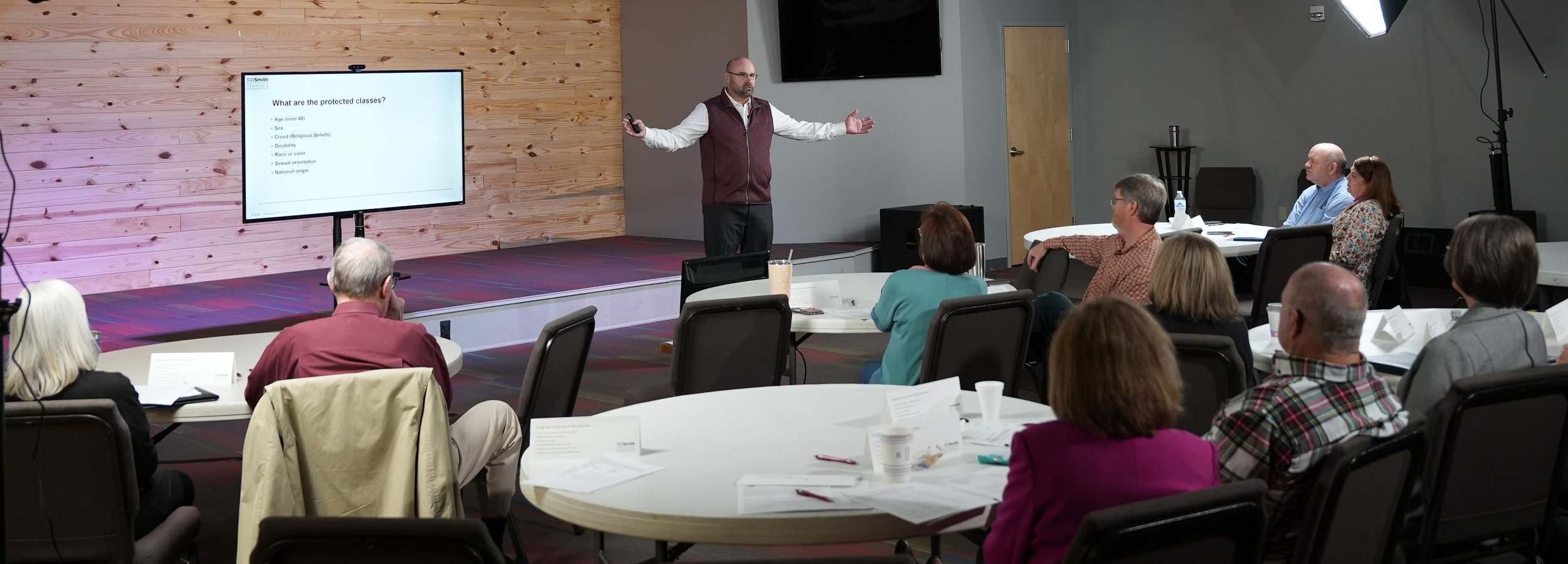 A man stands on a small stage giving a presentation to a seated audience in a conference room. The man is gesturing with open arms and is wearing glasses, a white shirt, and a maroon vest. A large screen behind him displays a slide titled 'What are the protected classes?' with bulleted points. The audience members are seated at round tables with papers, cups, and personal items in front of them.