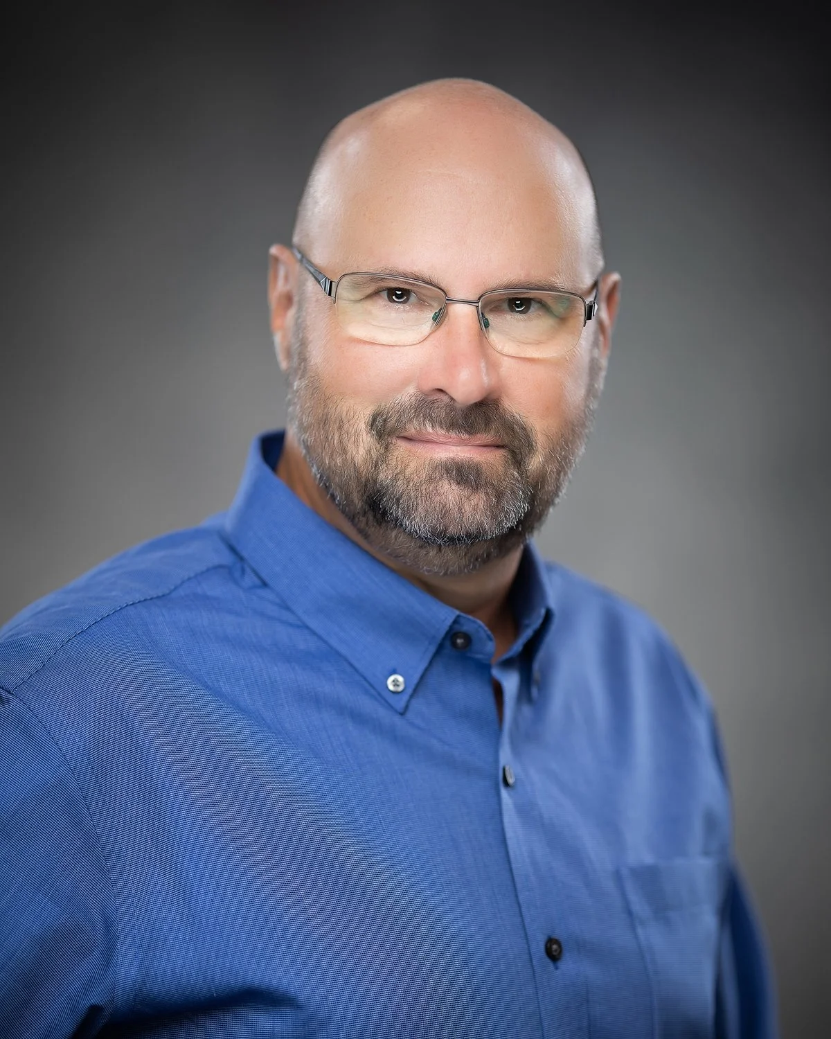 Headshot of a middle-aged man with a beard, wearing glasses and a blue collared shirt, against a dark gray background.