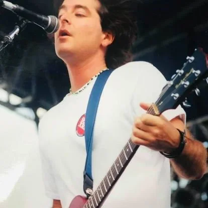 David Fagin singing into a microphone while playing an electric guitar during a live outdoor performance.