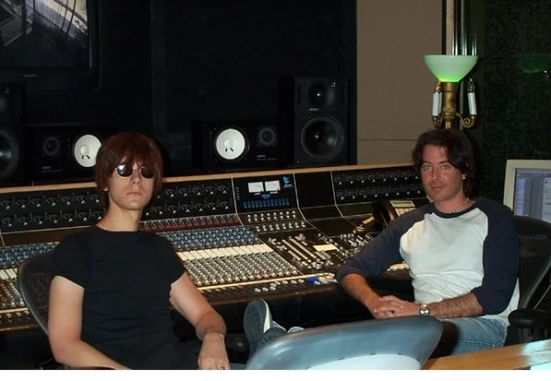 Two men sitting in a recording studio with audio mixing equipment and large speakers.
