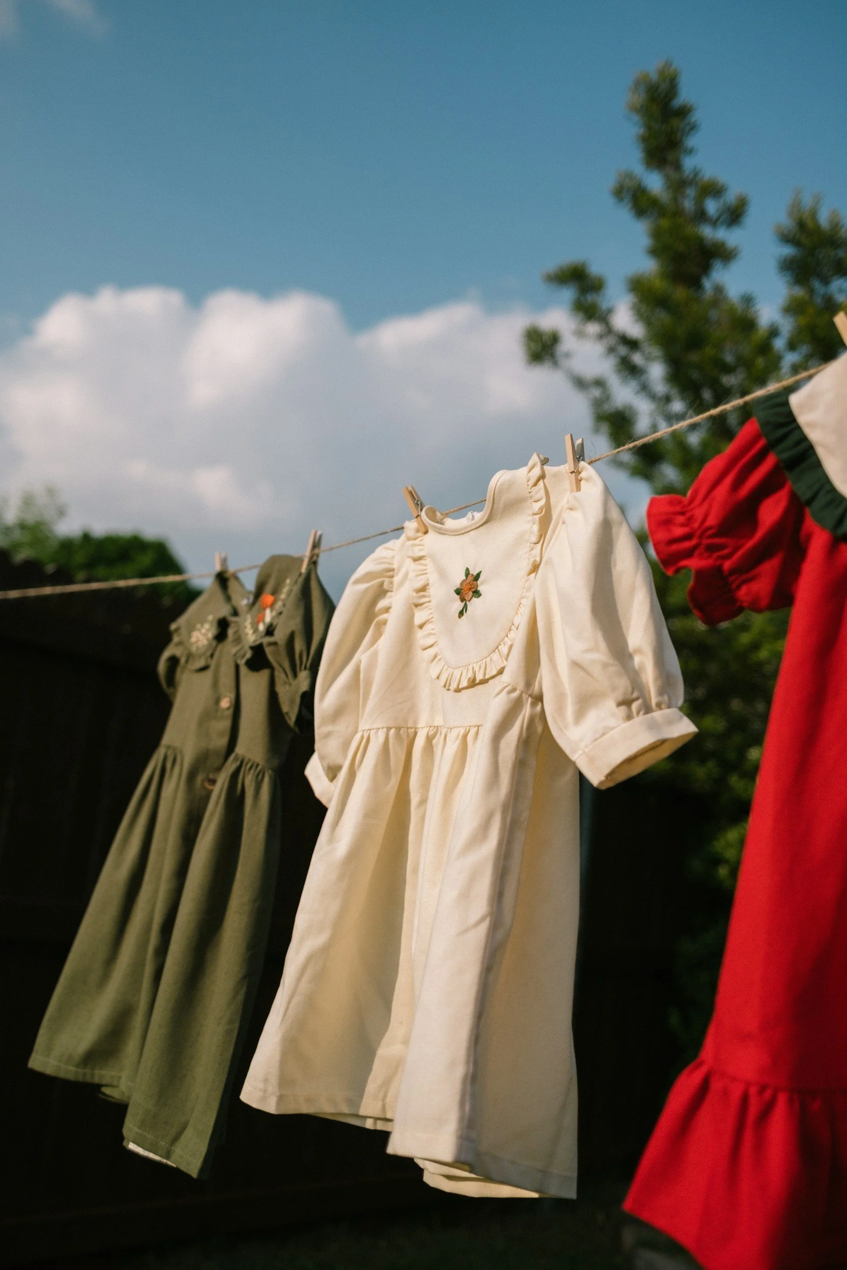 Clothes hanging on a clothesline, including a cream-colored dress with ruffled details and embroidered flowers, along with other garments in various colors.