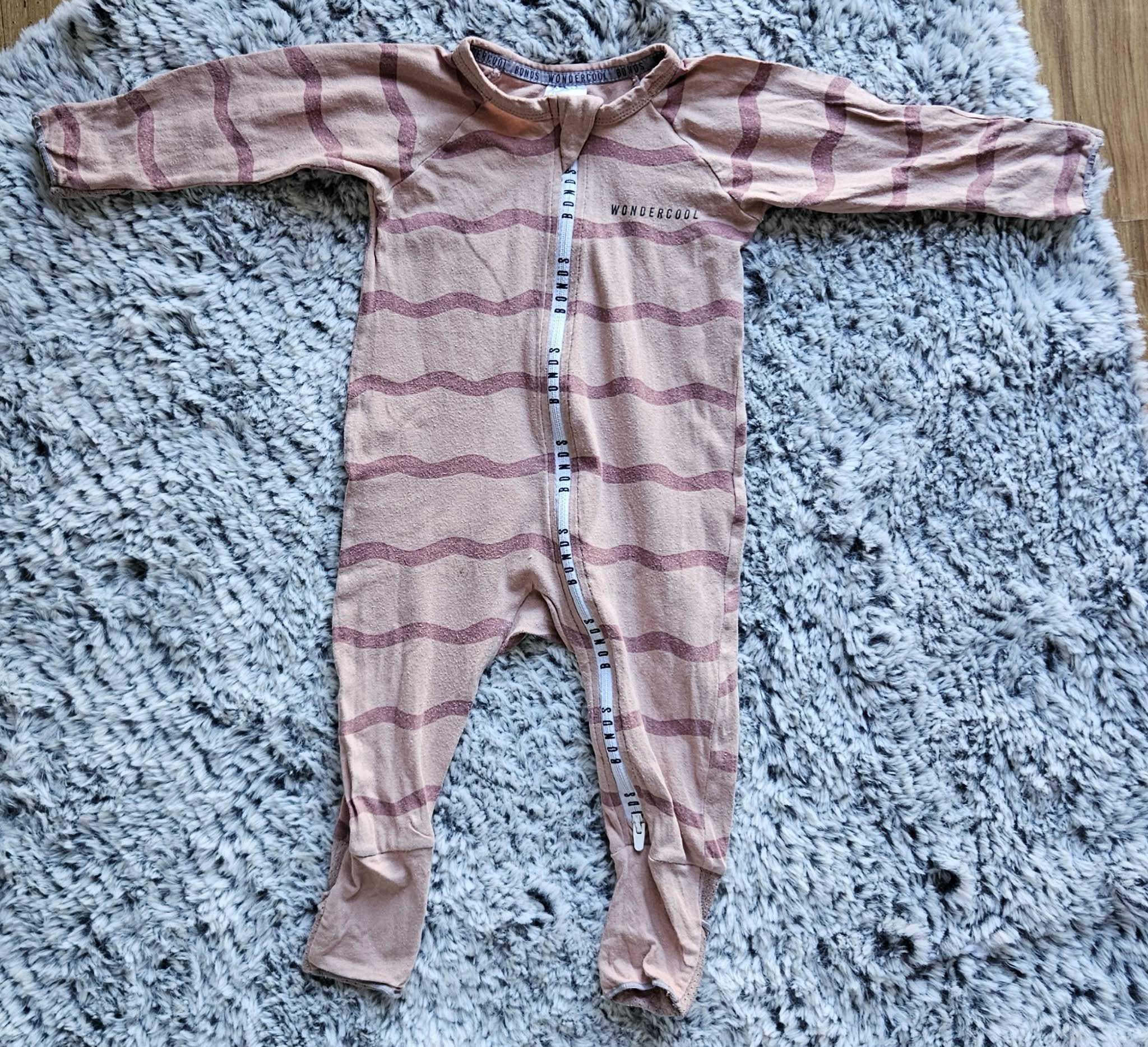 A pink and beige striped children's fleece jumpsuit with a zip front and a hood, laid flat on a fuzzy gray and white rug.