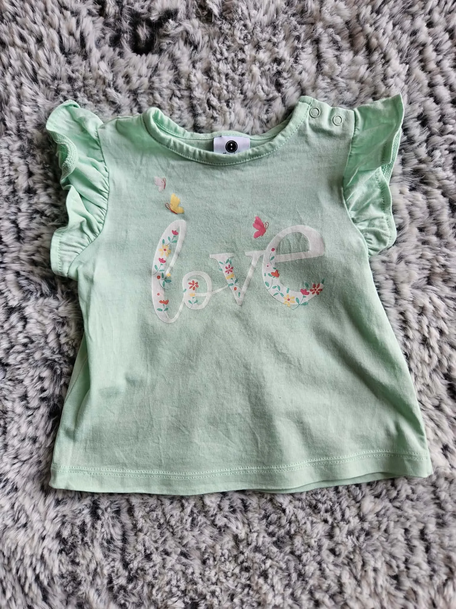 A light green baby onesie with ruffled sleeves and floral design on the front, laid on a gray and white furry surface.