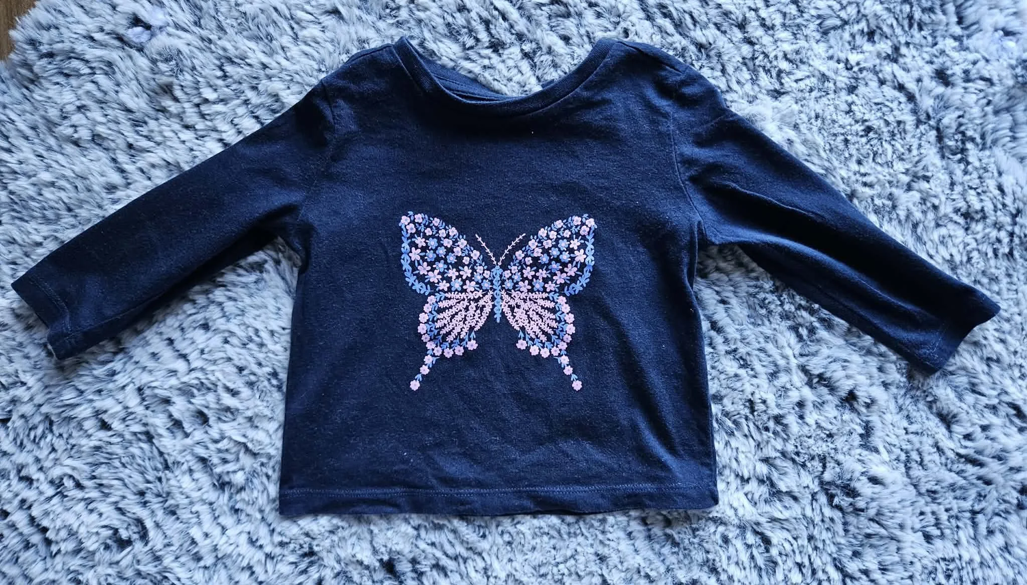 Dark blue long sleeve shirt with a pink and purple butterfly design made of small flowers, laid on a gray and white shaggy rug.