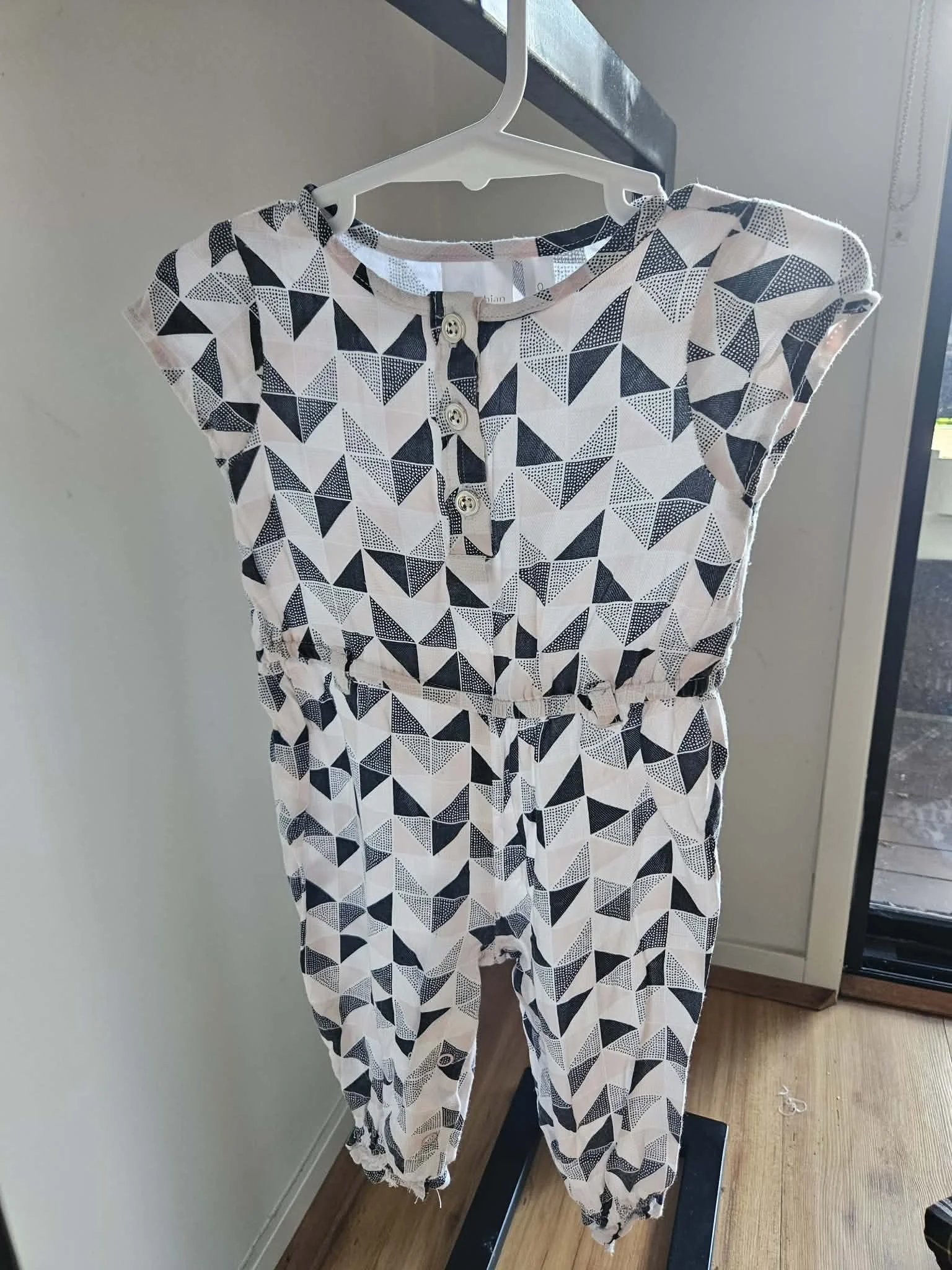 A white jumpsuit with a black geometric triangle pattern hangs on a white hanger in front of a mirror. The jumpsuit has a round neckline with a small slit fastened by four buttons and elastic cuffs at the ankles.