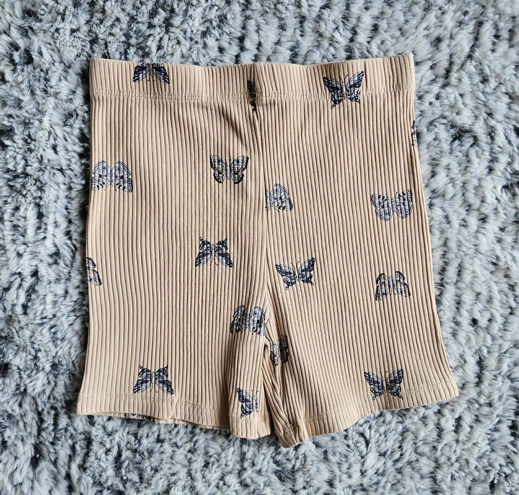 A pair of beige shorts with a butterfly pattern lies on a fluffy black and white textured surface.