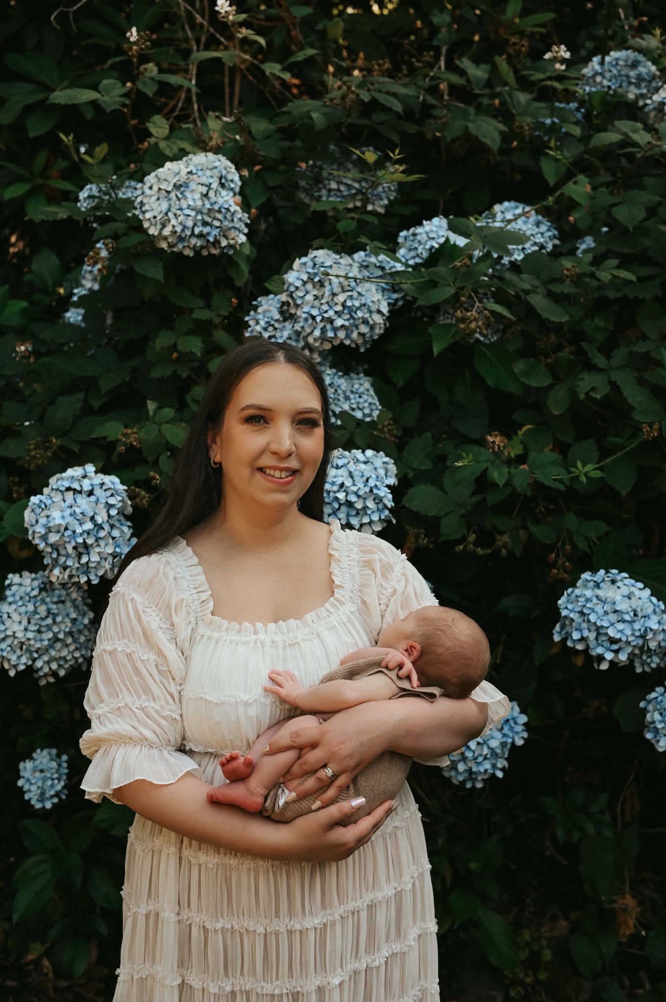A woman holding a baby in her arms, standing in front of a bush with blue hydrangea flowers. The woman has long dark hair and is wearing a light-colored dress. The woman is smiling at the camera, and the baby is resting peacefully in her arms.