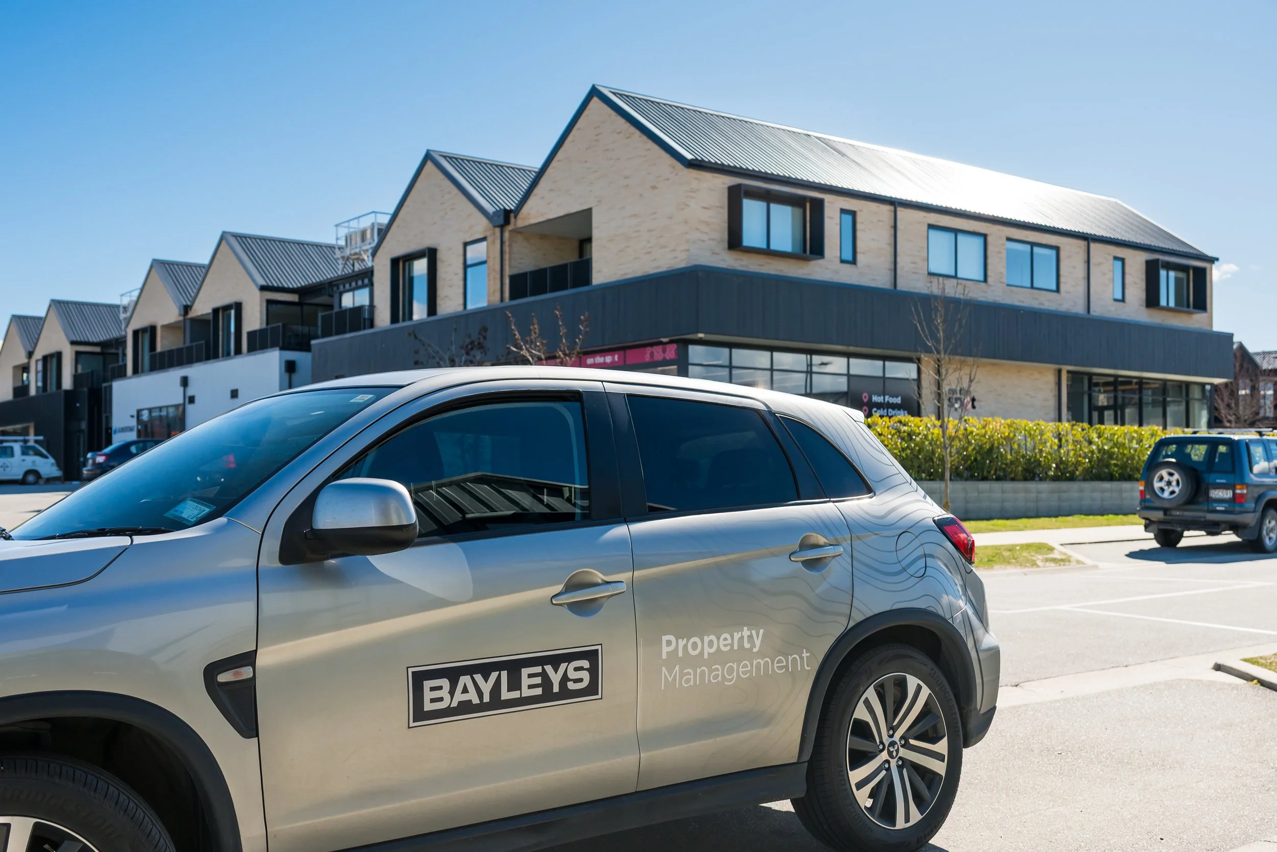 The Bayleys Commercial property management car in the parking lot of a modern commercial building in Wanaka with residential upper floors, set against a clear blue sky.