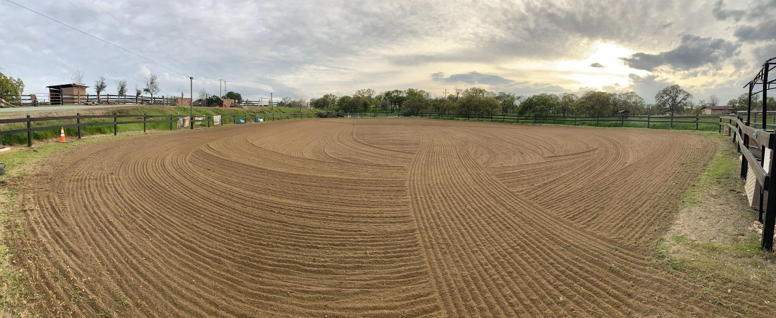 An empty dirt horse riding arena with freshly groomed ground and a black wooden fence surrounding it, under a cloudy sky with the sun partially visible.