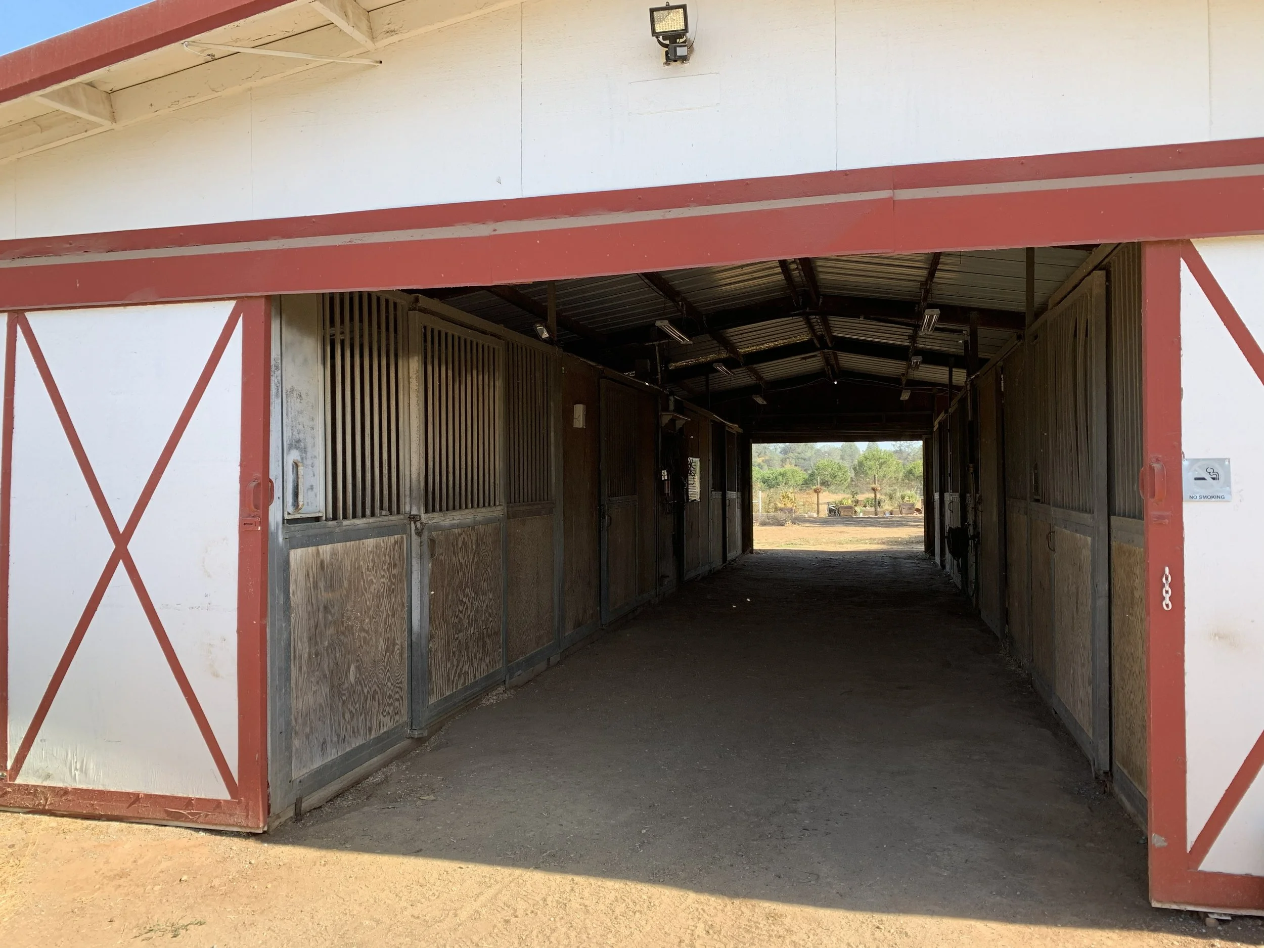 A horse stable with wooden stalls under a red and white roof, situated outdoors with trees in the background.