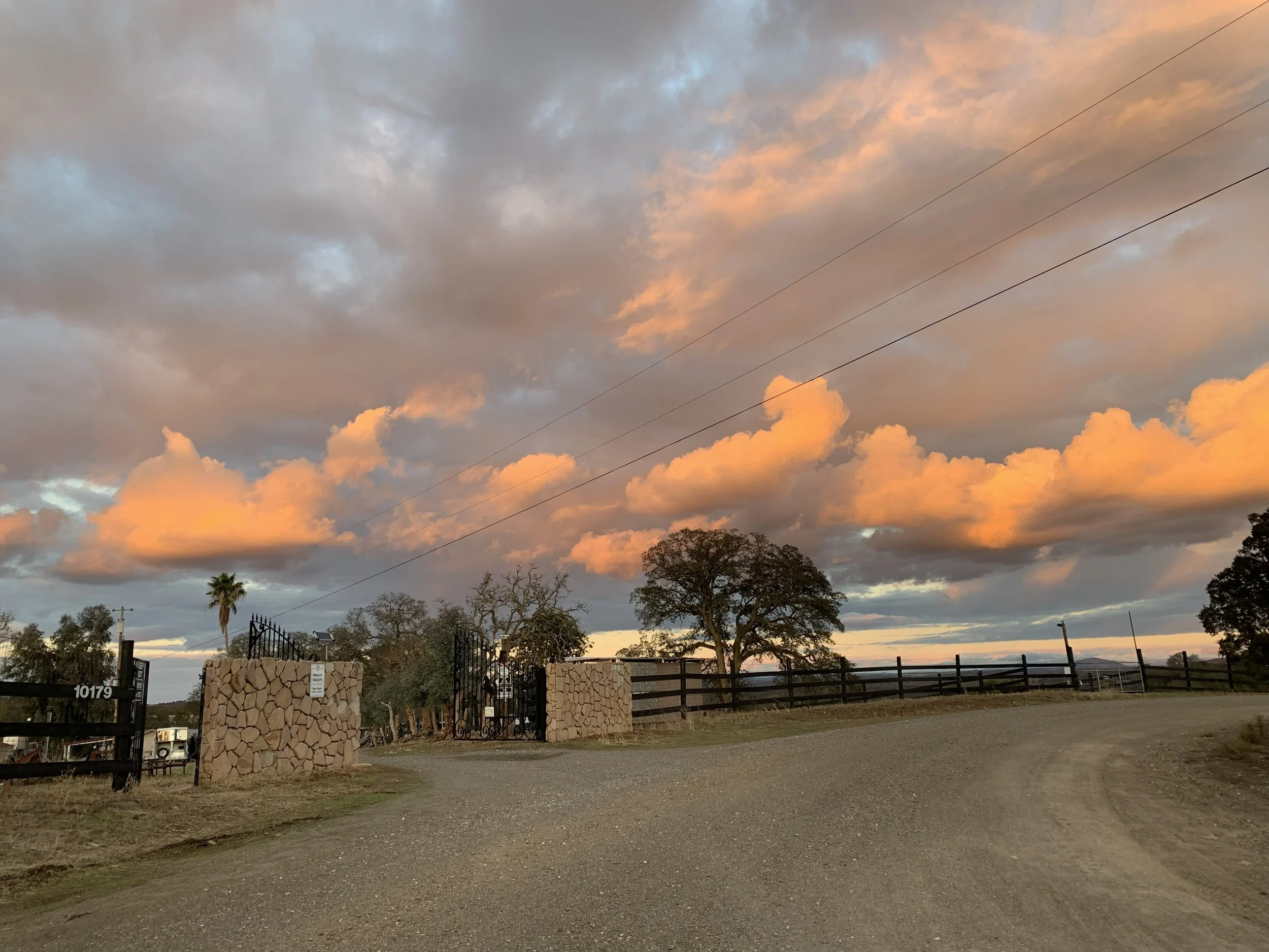 A rural scene at sunset with a partly cloudy sky, trees, a stone and metal gate, fencing, and a gravel driveway.