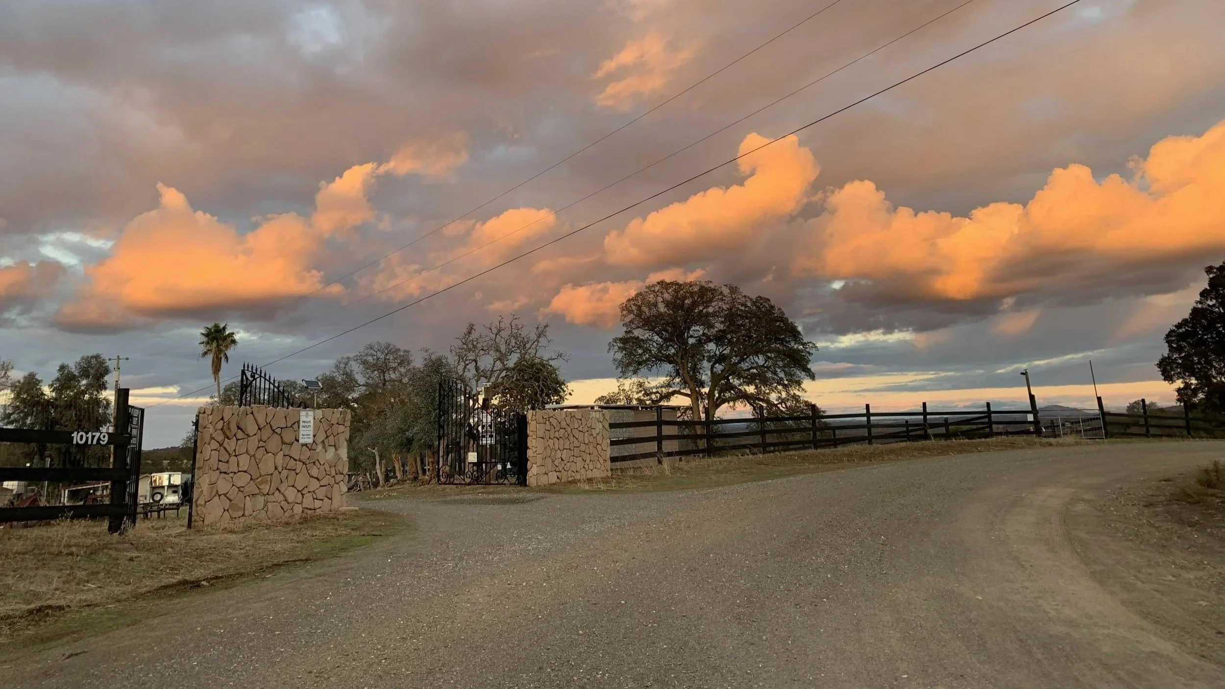 A dirt entrance road leading to a gated property with stone pillars, wooden fence and trees, under a colorful sky with orange and gray clouds at sunset.