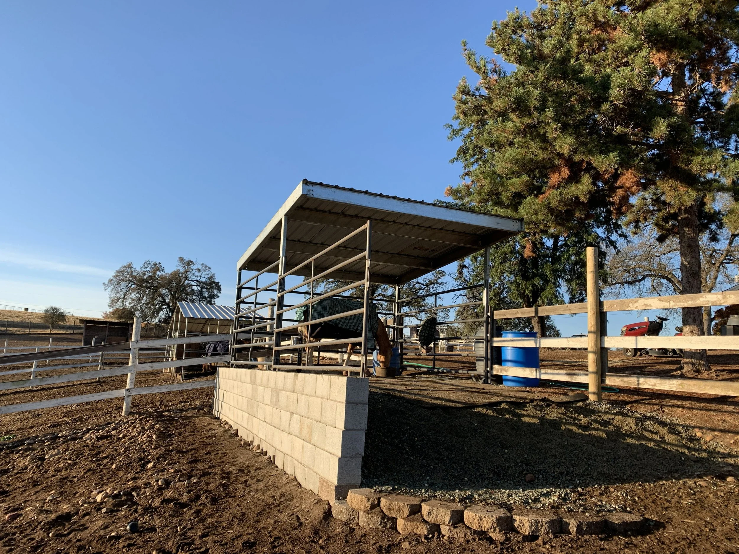 A partially constructed outdoor structure with a cinder block wall, metal framework, and a corrugated metal roof in a rural setting with trees and open land.