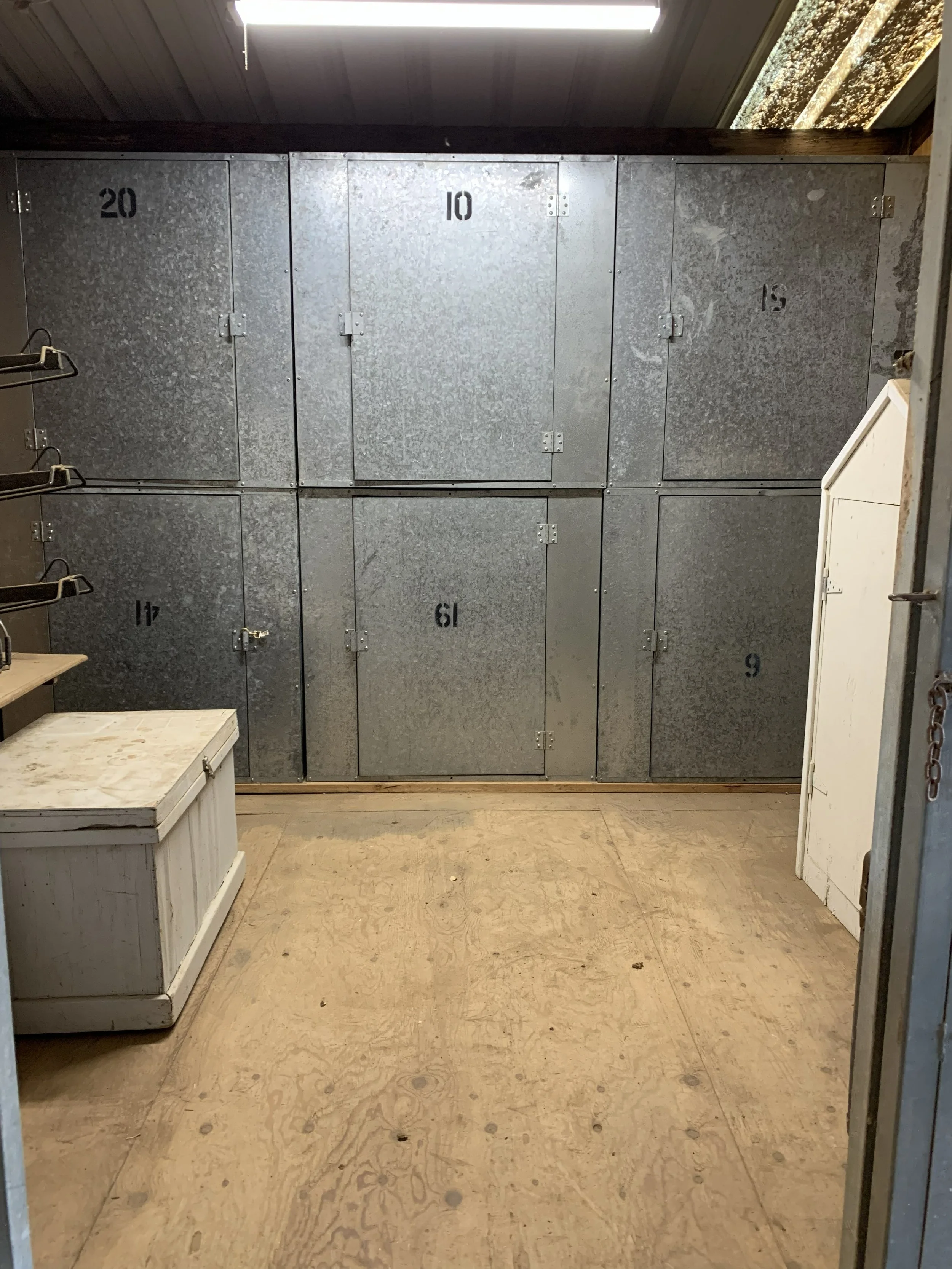 A room with metal lockers numbered 20, 10, 15, 14, 13, and 9, with a white wooden chest and a small white wooden cabinet in the foreground, and a fluorescent light fixture on the ceiling.
