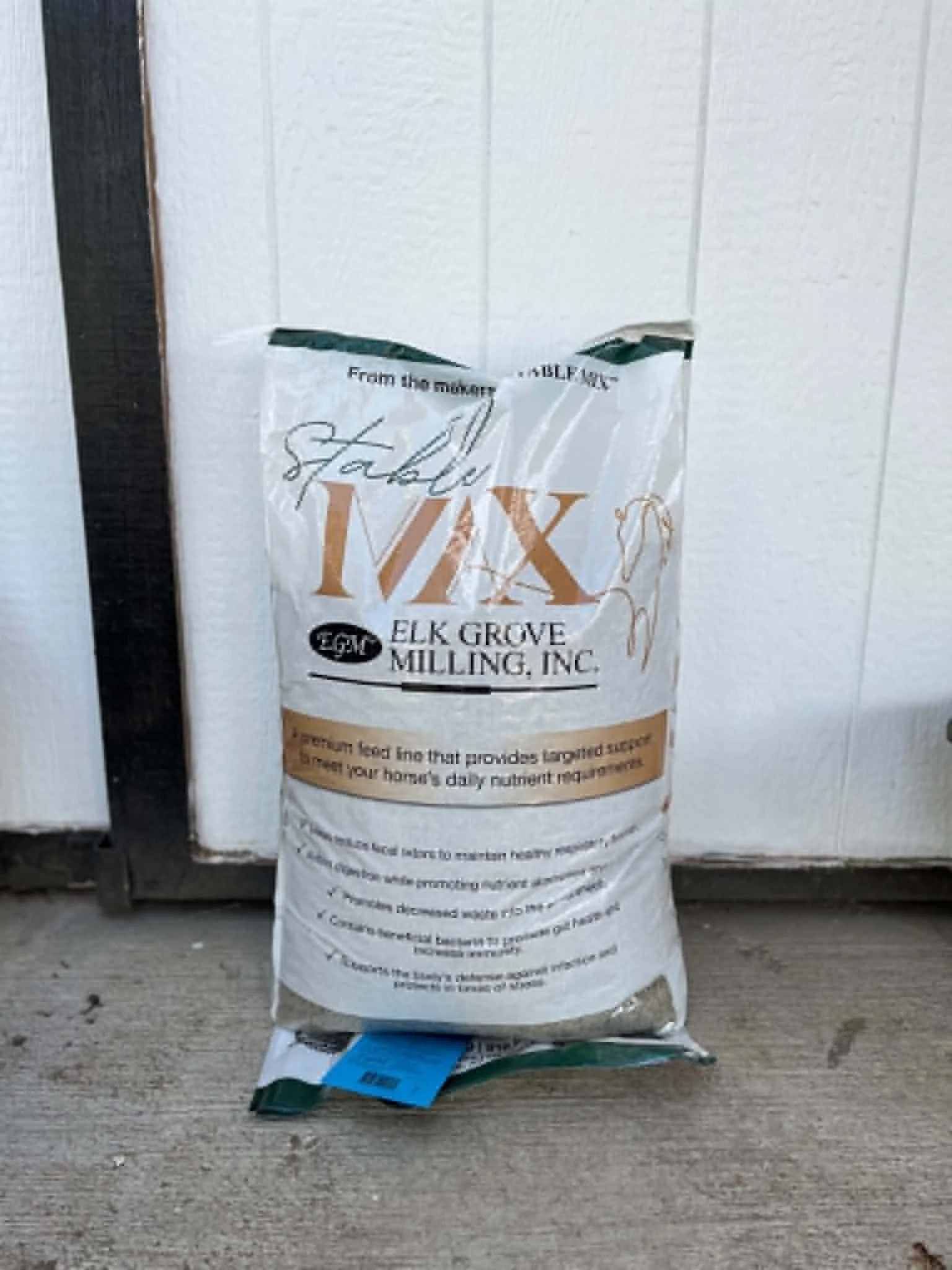 A bag of Flakkie IVX chicken feed from Elk Grove Milling, Inc., placed on a concrete surface near a white wooden wall.
