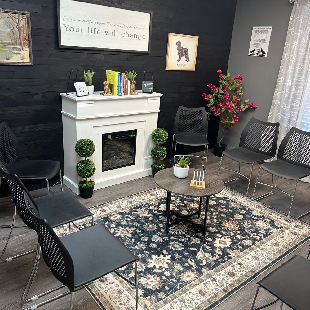 A cozy waiting room with black walls, a white fireplace, and black chairs arranged in a semi-circle. There is a small round wooden table with a white plant and a candle holder. Decor includes framed dog artwork, a large sign that reads "Your life will change," potted plants, pink flowers, and a patterned rug. Curtain partially covers a window.