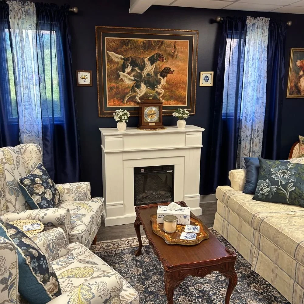 A living room with dark navy walls and blue curtains. There is a white fireplace with a clock and two vases with white flowers on top. Above the fireplace, there is a painting of two dogs running in a field. The room has floral-patterned sofas with cushions and a wooden coffee table with a tissue box, candles, and coasters on it. There is a patterned rug on the floor.