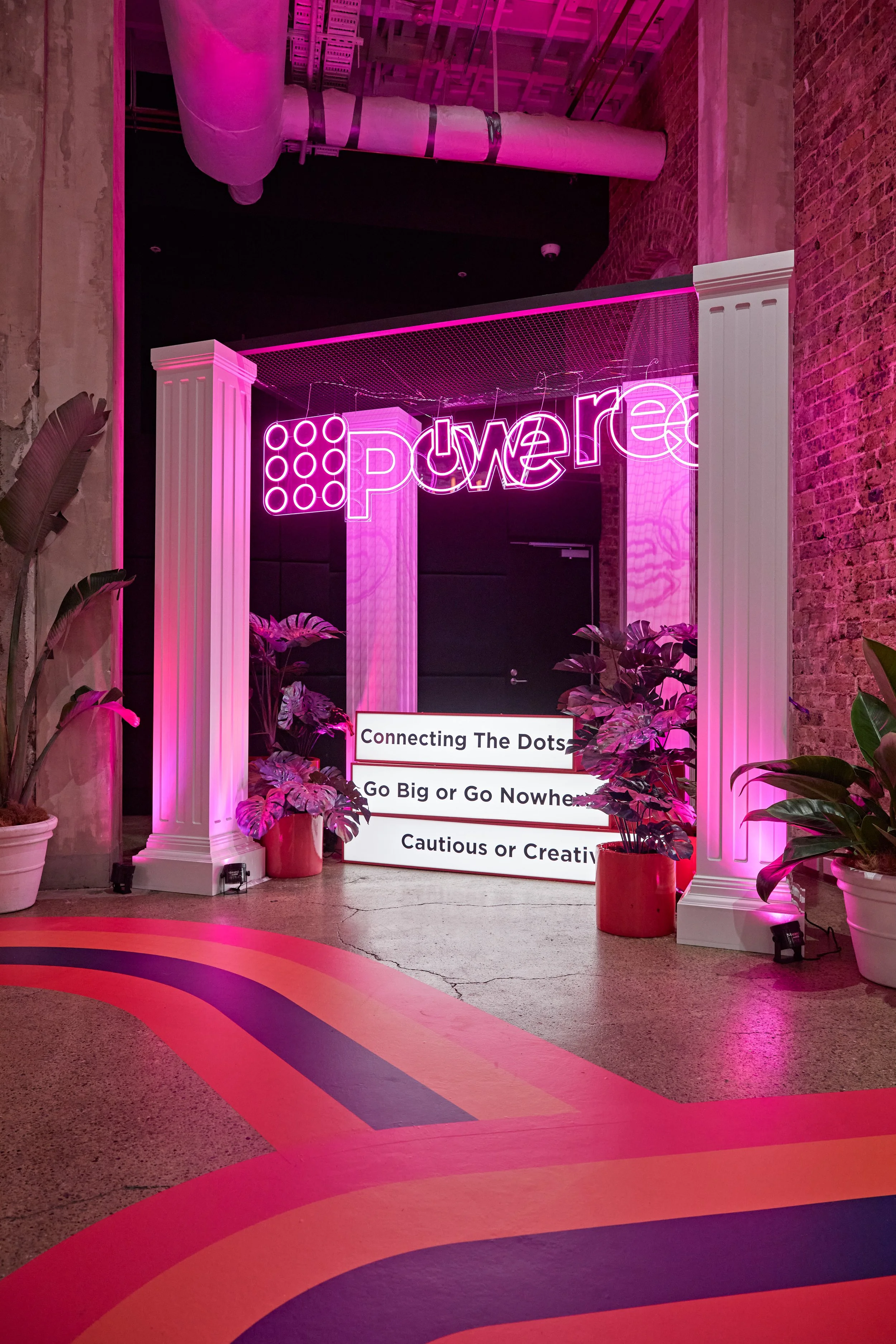 Pink neon sign reading 'Power' hangs above pink and white columns with potted plants in red and white pots. A colorful rainbow pattern decorates the floor, and there is a white sign with black text about connecting dots, going big or no, and being cautious or creative. The setting appears to be an indoor event space or gallery with an industrial feel, exposed brick walls, and ductwork on the ceiling.
