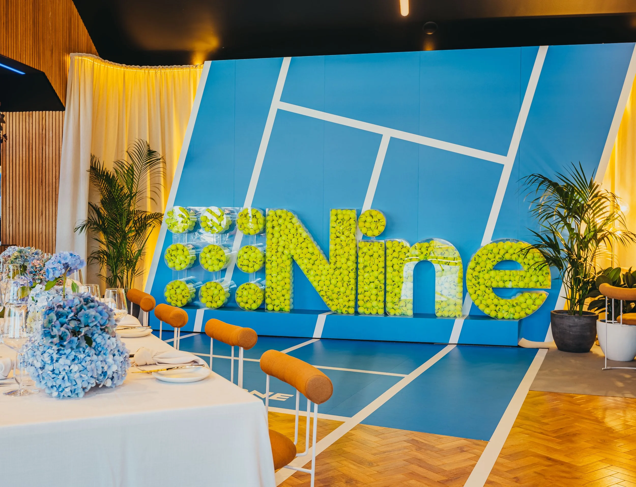 Indoor tennis-themed event space featuring a large blue wall with a tennis court design and compartments filled with green tennis balls. The room has decor including potted plants, a long table with flower arrangements, and orange chairs.