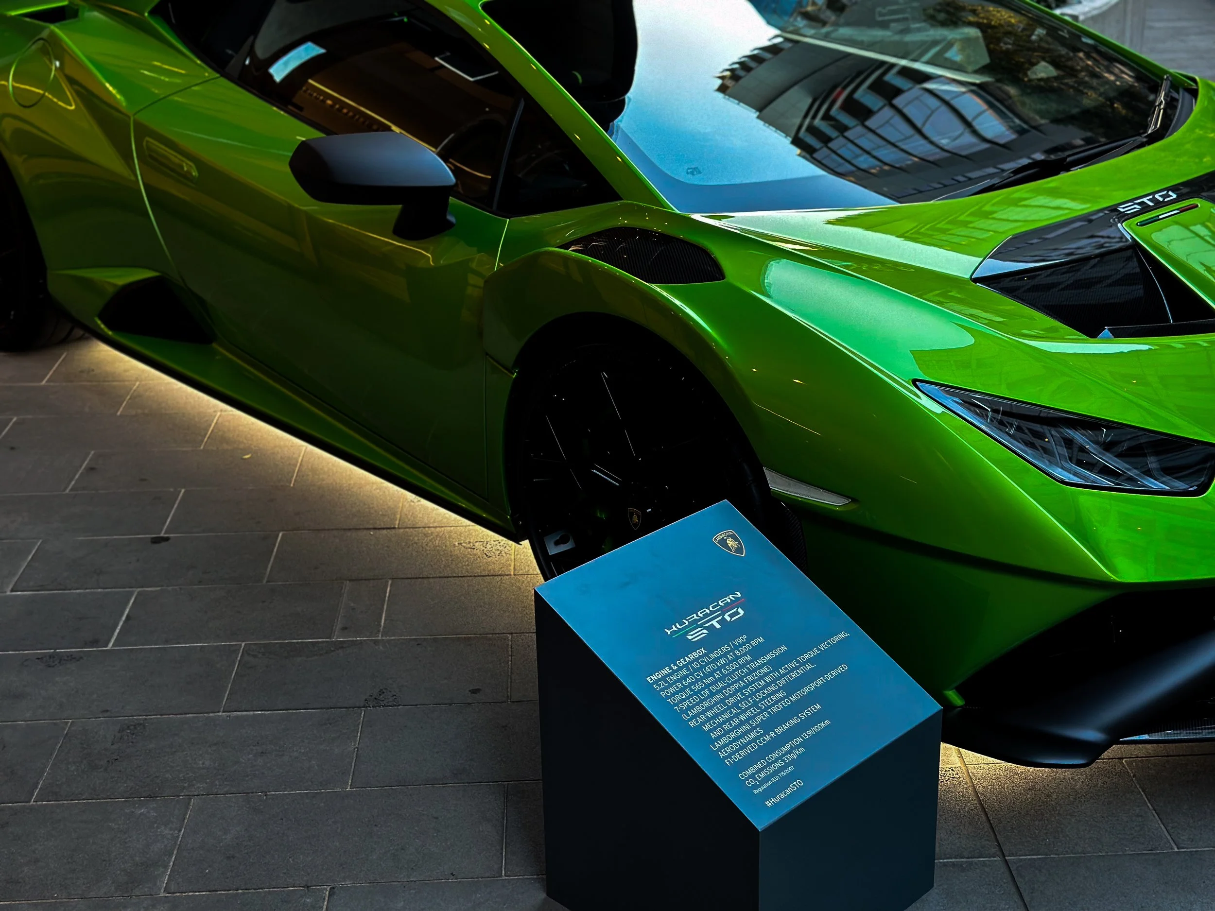 A bright green luxury sports car displayed indoors with a blue informational sign in front of it.