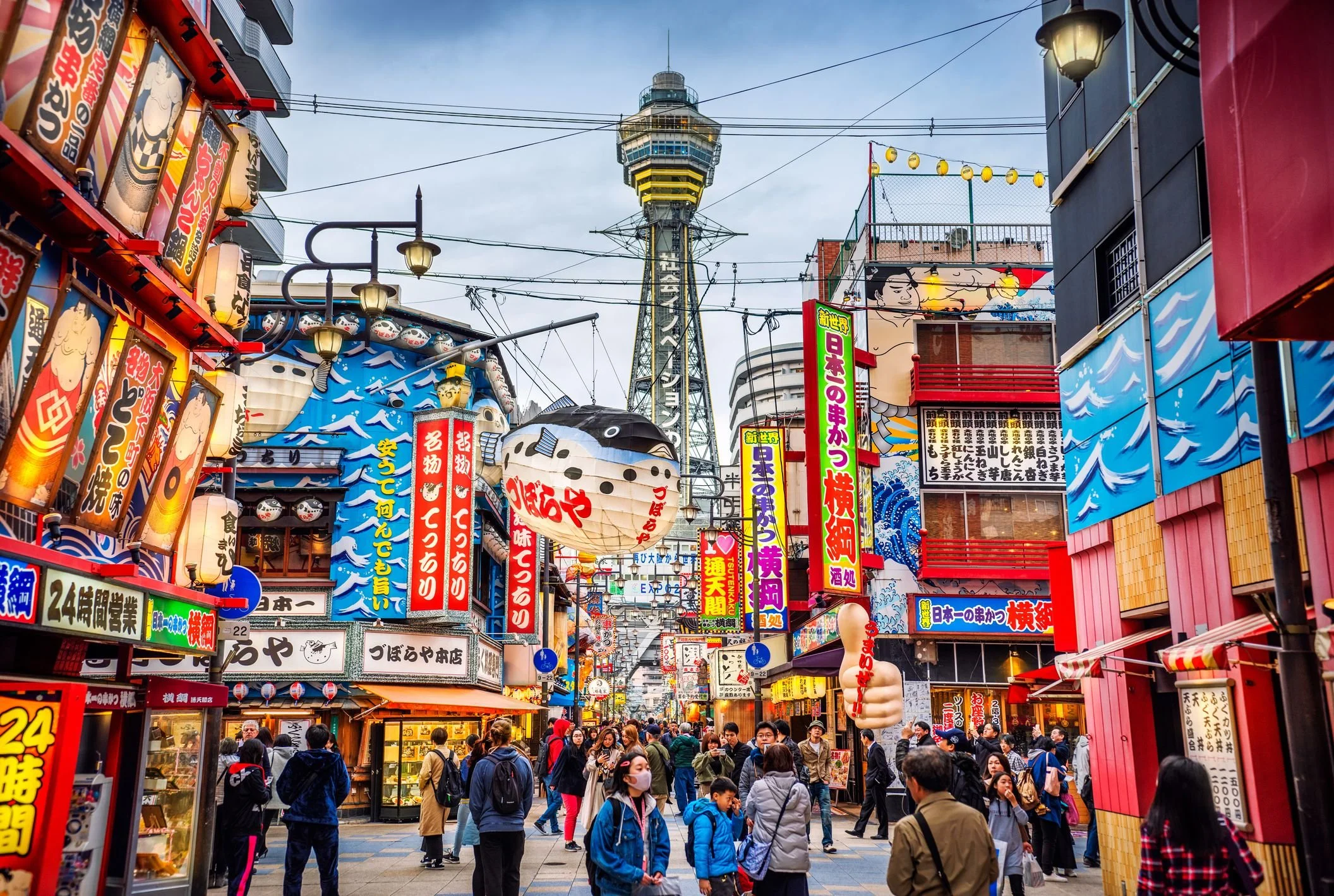A busy street scene in Japan with numerous colorful signs, lanterns, and a tall tower in the background. Crowds of people walk along the street, many wearing face masks.