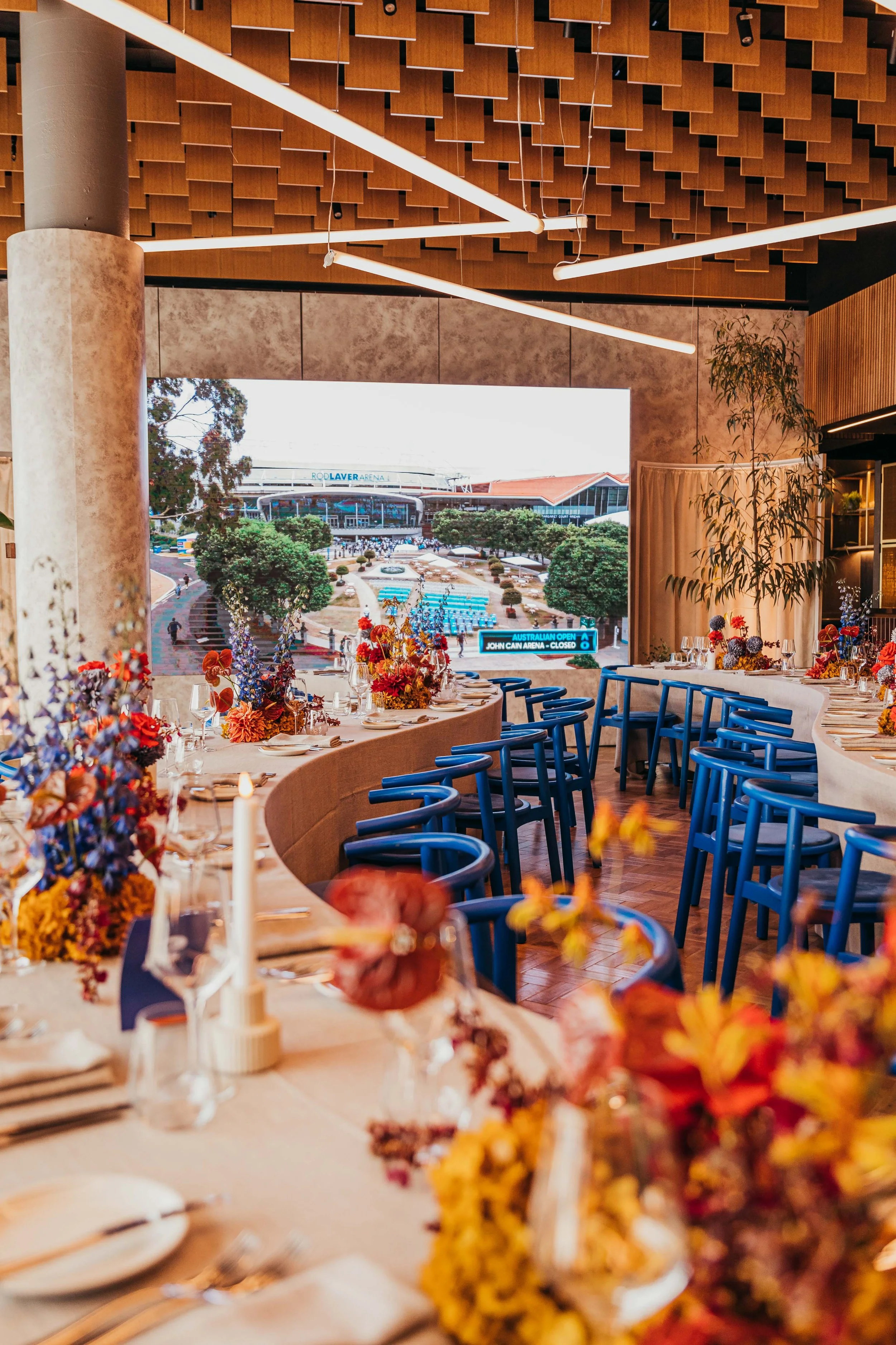 Indoor dining area with table settings and floral centerpieces, overlooking a view of the outdoor area with trees, a pathway, and a stadium in the background.